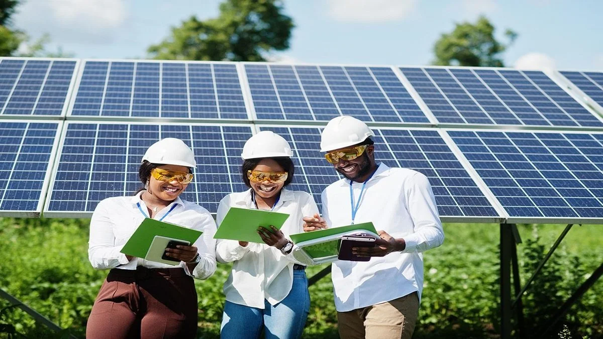 Three diverse energy professionals wearing white helmets and safety glasses inspecting solar panels outdoors, holding green folders and documentation.