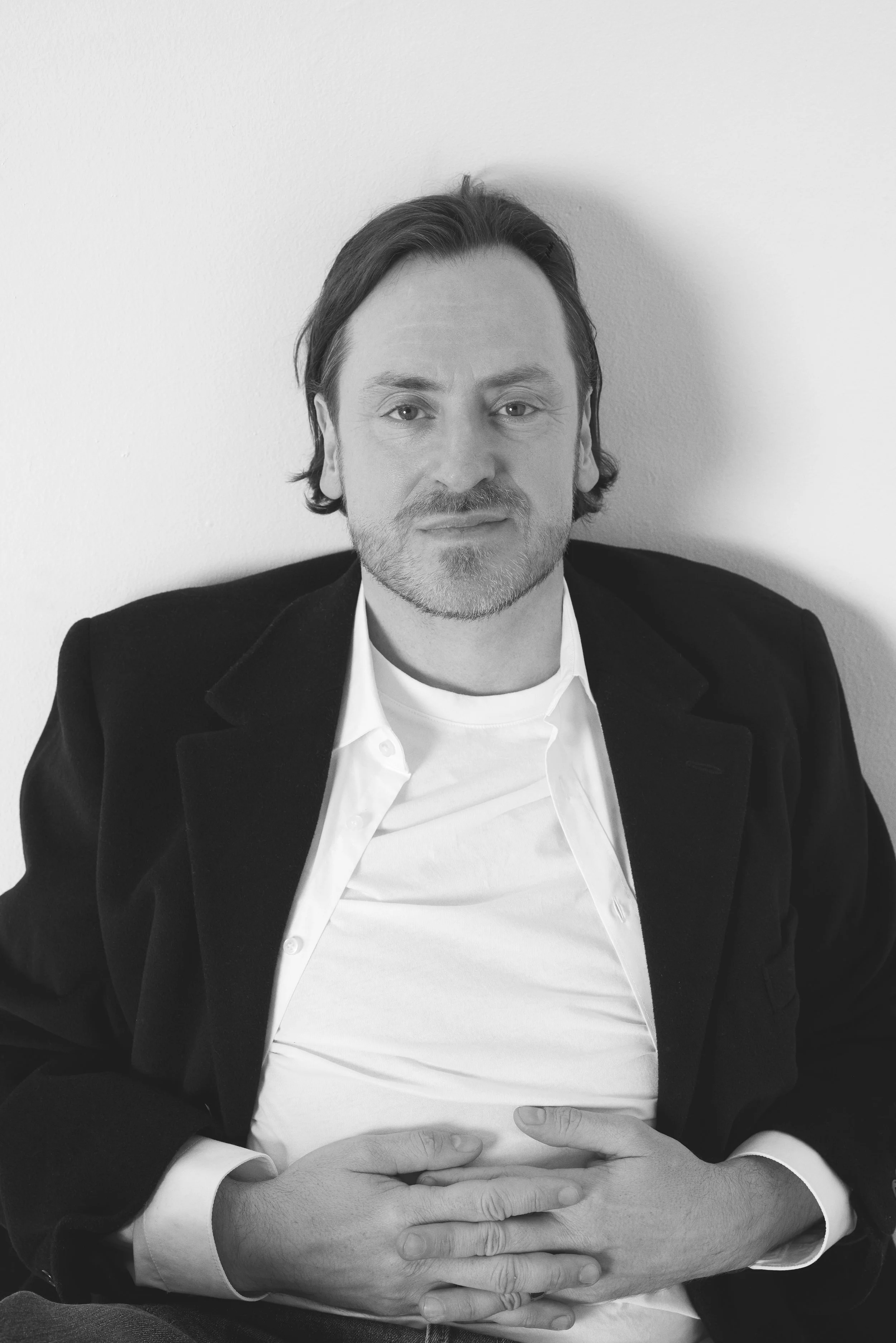 Black and white portrait of a man with dark hair and facial hair, wearing a white shirt and a dark blazer, with hands clasped on his lap, sitting against a white wall.