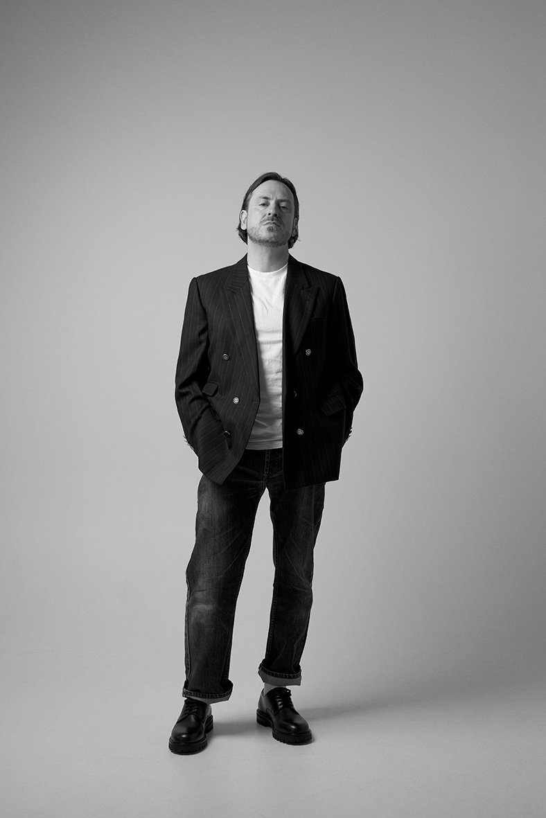 A man with medium-length hair and a beard, wearing a dark pinstripe blazer over a white shirt, with jeans and black boots, standing with hands in pockets against a plain light gray background.