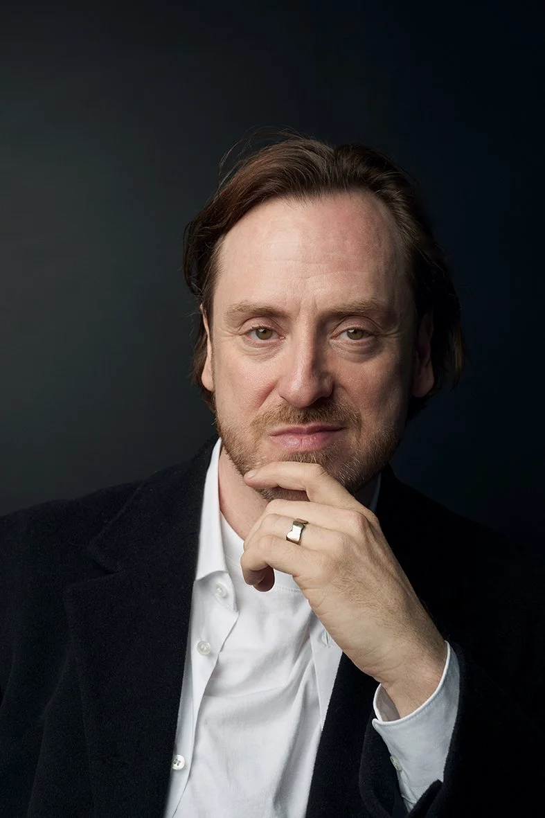A man with brown hair and a beard holding his chin with his right hand, wearing a black blazer and white shirt, against a dark background.
