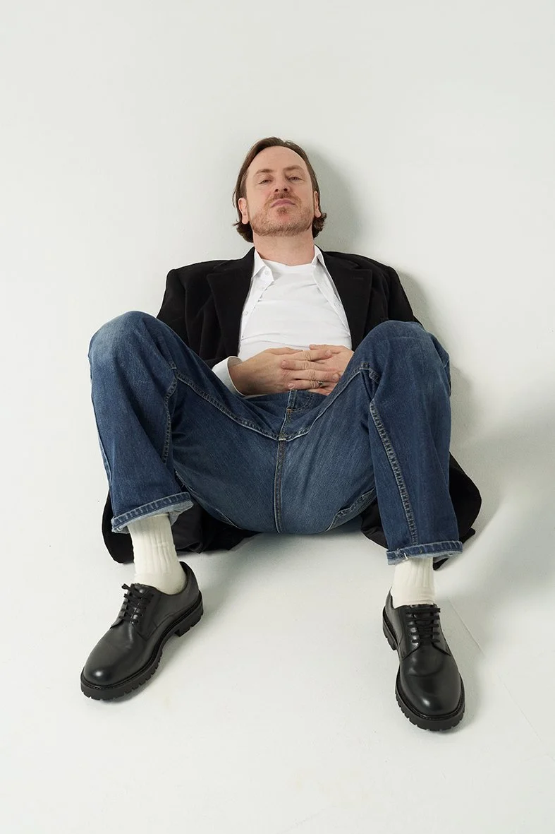 A man sitting on the floor with his legs spread and his hands on his stomach, leaning against a white wall. He is wearing a black blazer, a white shirt, blue jeans, white socks, and black shoes.