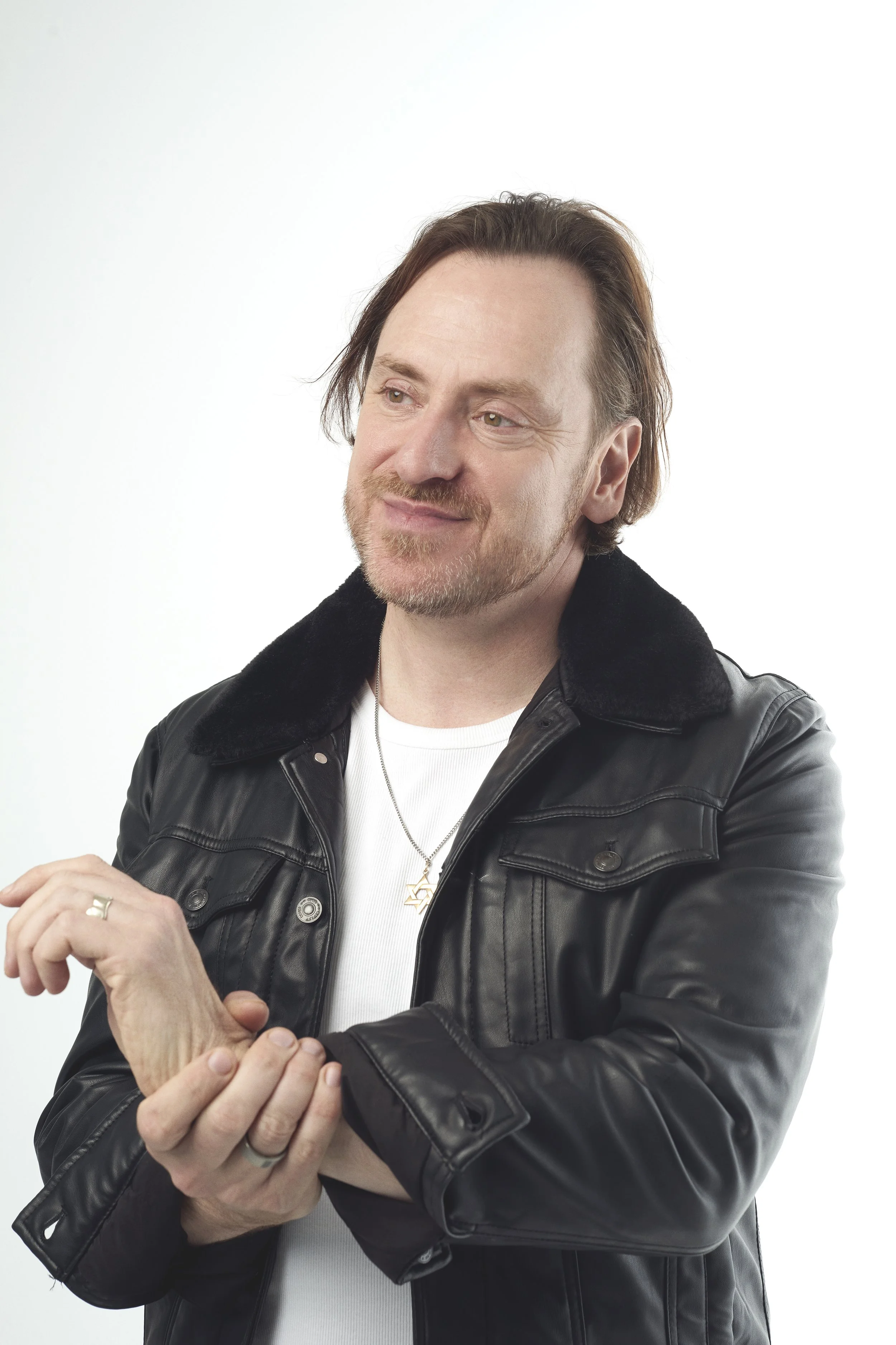 A man with brown hair, a beard, wearing a black leather jacket with a fur collar, a white shirt, and a star-shaped necklace, is smiling and looking to his left. He is holding his right wrist with his left hand, and the background is plain white.