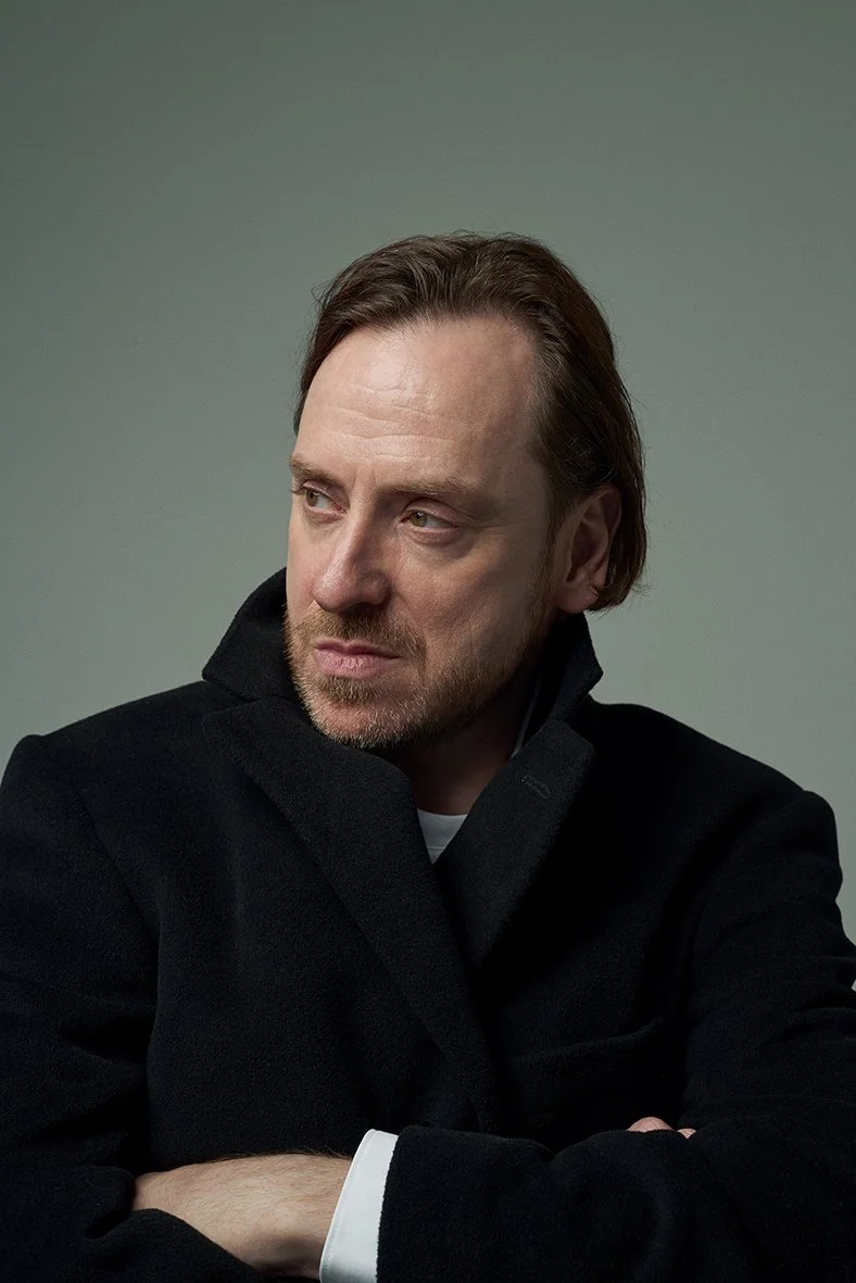 Headshot of a man with brown hair and a beard wearing a black coat, looking to the side against a gray background.