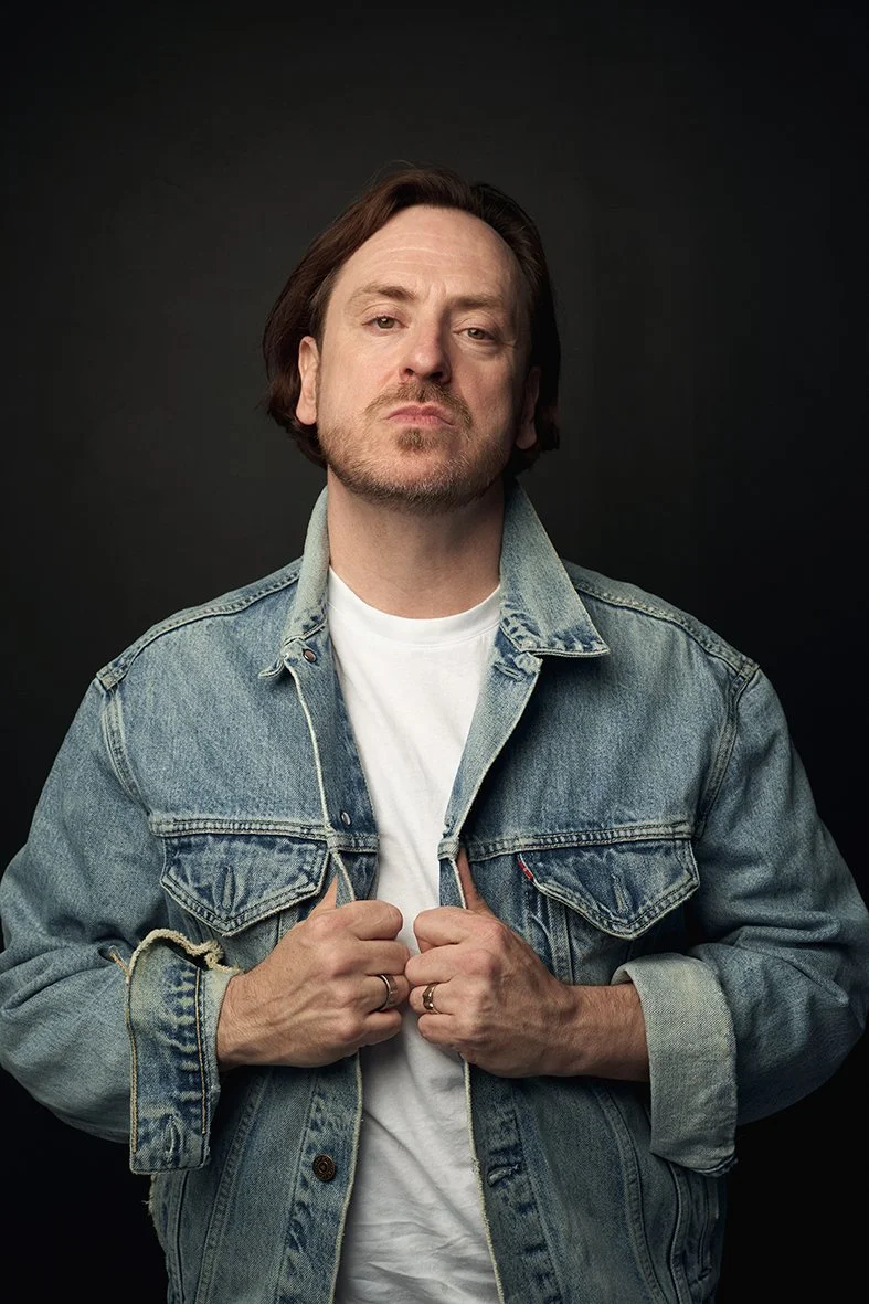 A man with medium-length brown hair and a beard, wearing a denim jacket over a white T-shirt, looking confidently at the camera against a dark background.