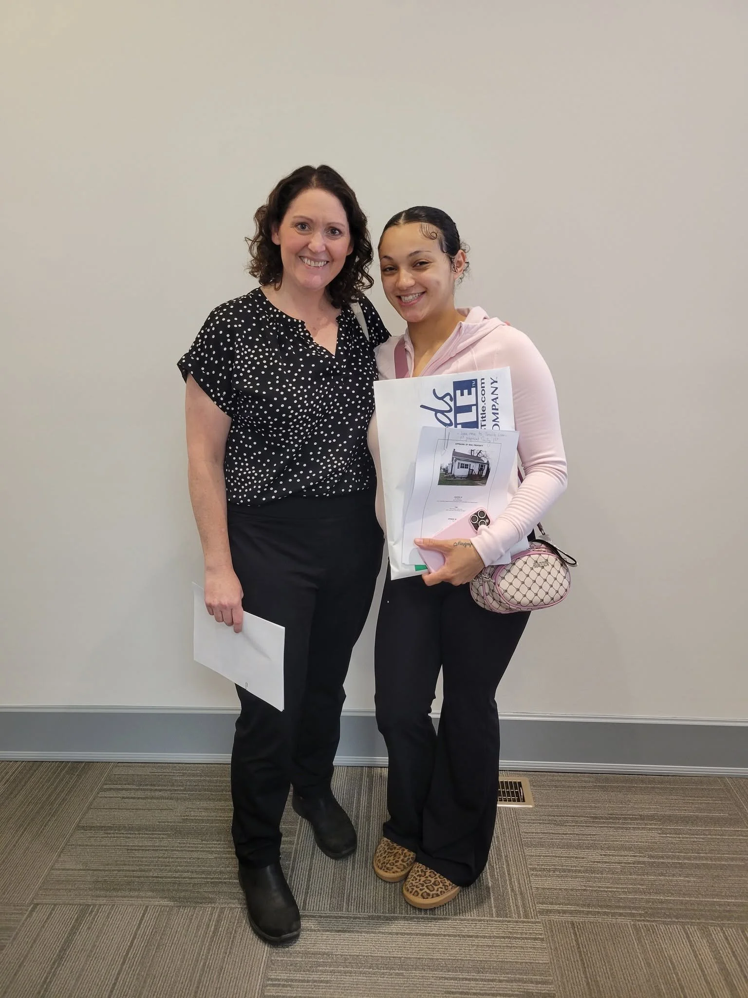 Two women standing together, smiling, holding papers and a brochure, dressed casually, against a plain wall. One is wearing a black polka dot top and black pants, the other in a pink hoodie and patterned slippers.