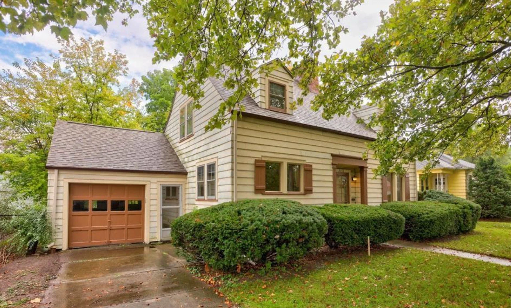 A two-story suburban house with cream-colored siding, brown window shutters, a brown garage door, and a brown front door. The house is surrounded by well-maintained bushes and trees, with a concrete driveway leading to the garage.