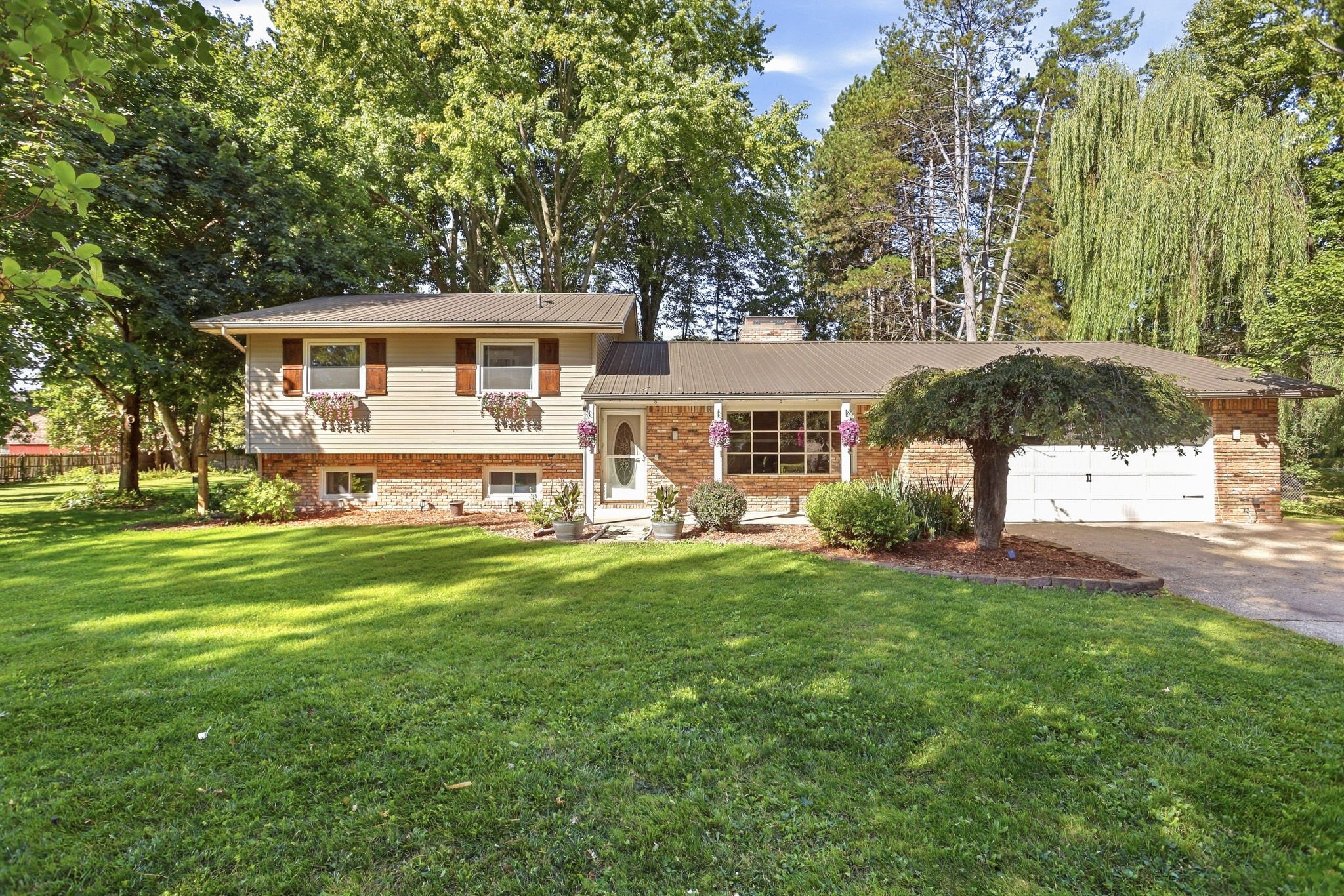 A suburban house with a well-maintained front yard, green lawn, and large trees providing shade. The house has a brick and beige siding exterior with hanging flower baskets near the windows and a white garage door. A paved driveway runs along the fro