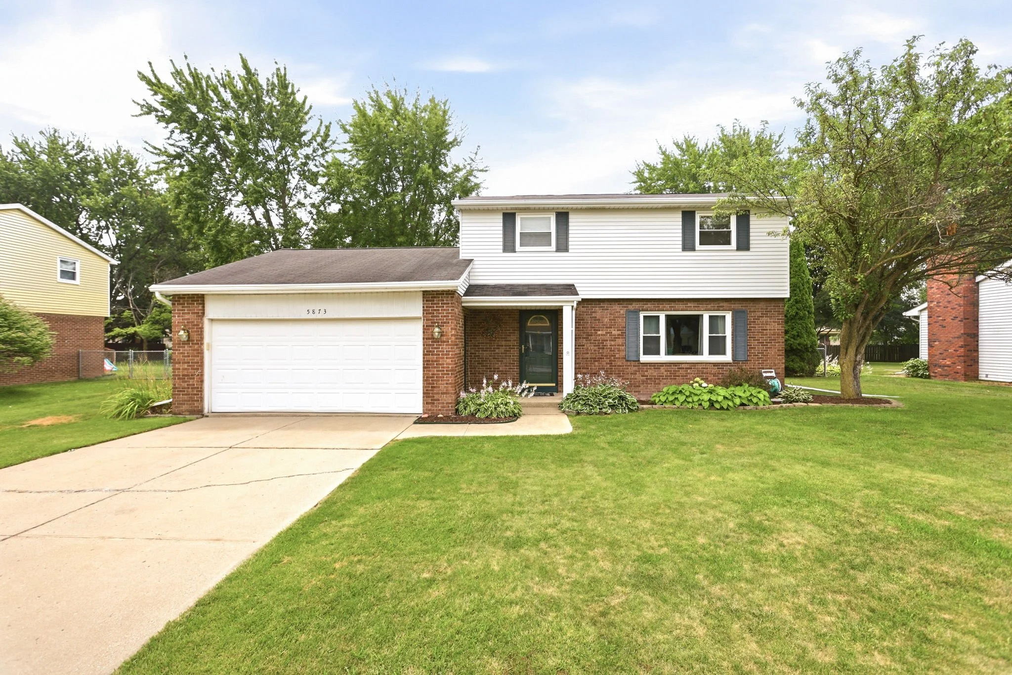 A two-story house with a brick lower level and white siding upper level, a two-car garage, a green front door, and a manicured lawn with flower beds and trees.