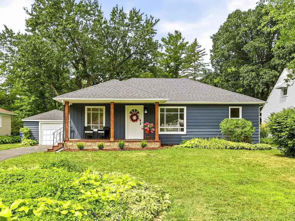 Front view of a blue house with a gray roof, small front porch with two black chairs, white front door with a red wreath, and a flowerpot with pink flowers. Green lawn and trees in the background.