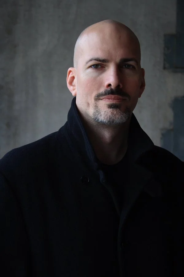 A bald man with a goatee and mustache, wearing a dark coat, standing against a plain, gray background.