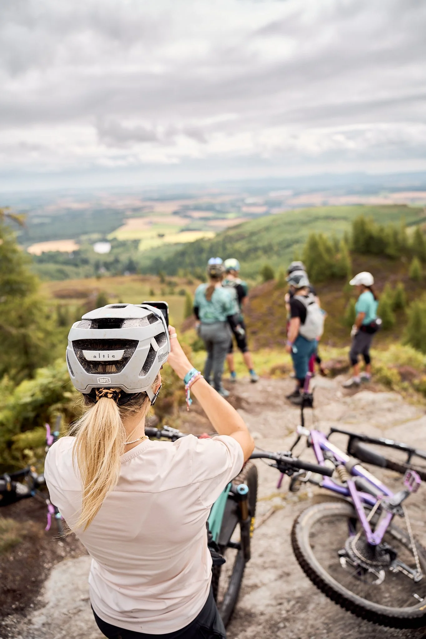 Back view of a woman in a beige shirt and a helmet taking a photo with her phone during a mountain biking trip, with bicycles and other people in the background on a dirt trail surrounded by greenery and a vast landscape under cloudy skies.