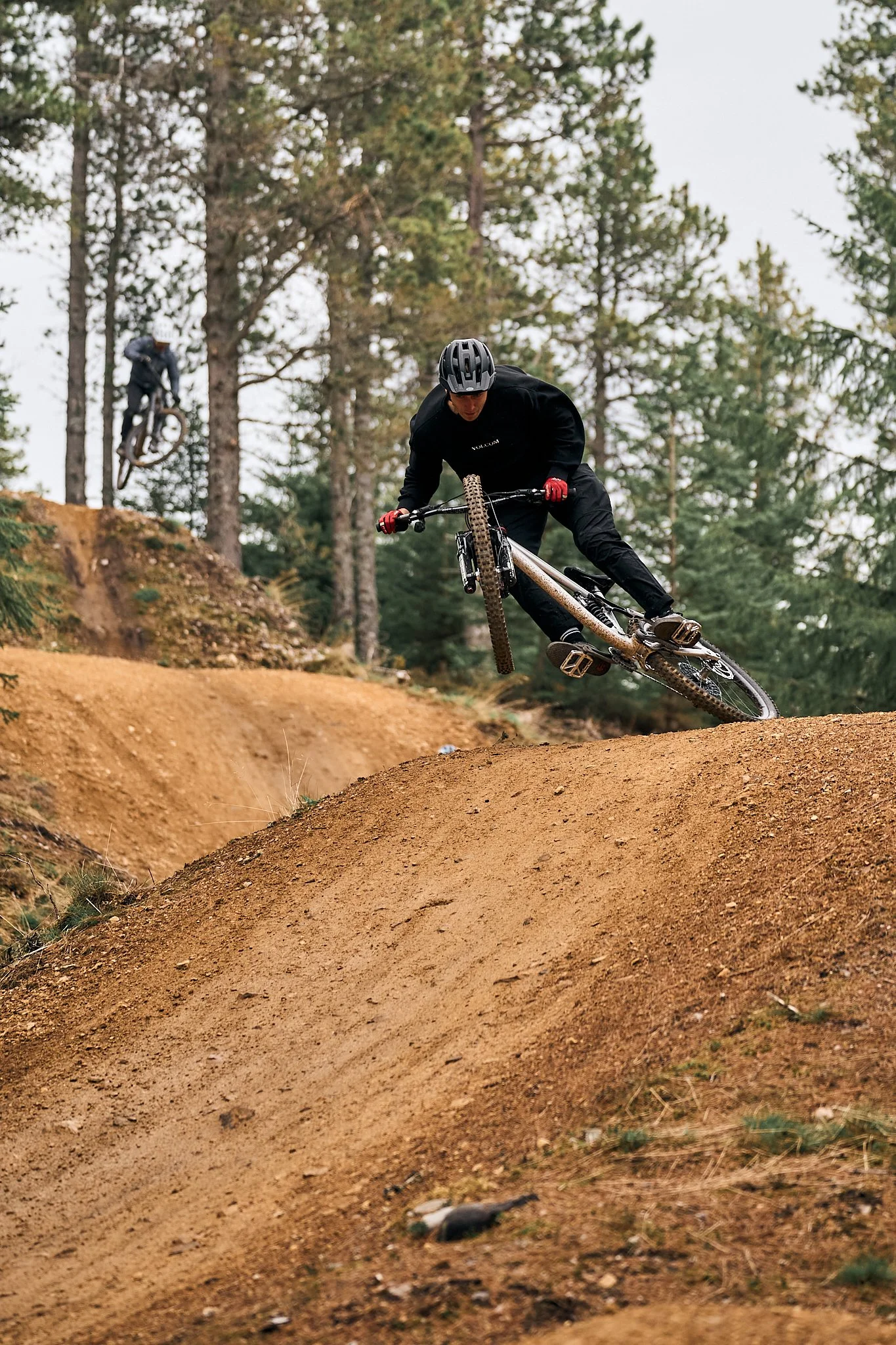 Two mountain bikers riding on a dirt trail in a forested area, one in the foreground and the other in the background, both wearing helmets and protective gear.