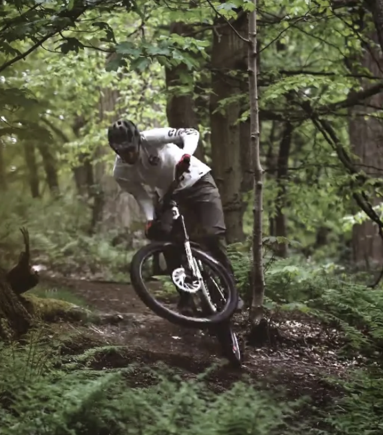 Mountain biker wearing a helmet and white jersey riding through a forest trail, leaning sharply as they navigate around a tree.