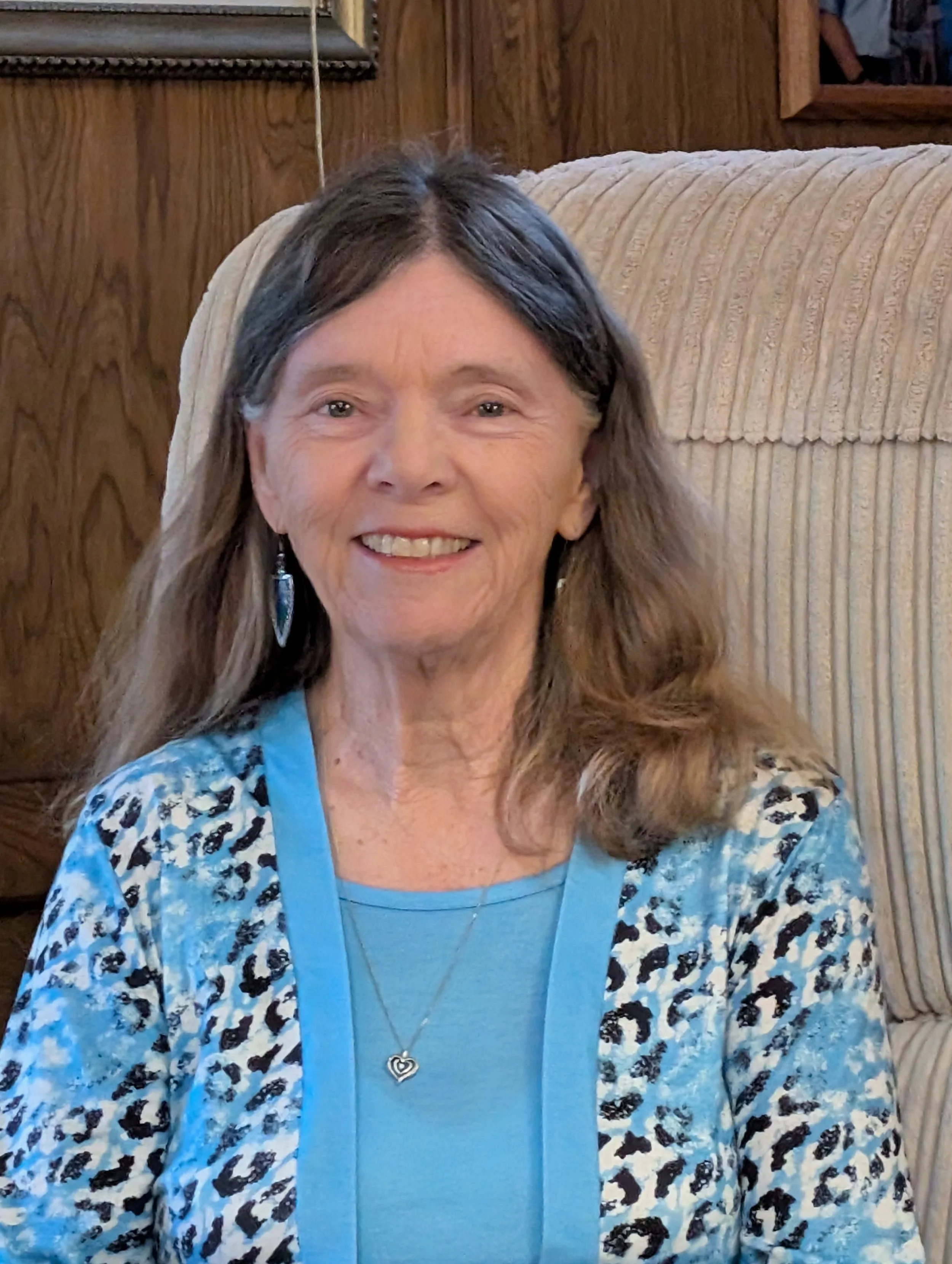 Sheila E. Humphrey. A smiling woman with long, wavy brown hair sitting on a beige upholstered chair with a wooden wall and mirror in the background. She wears a blue top with a heart necklace.