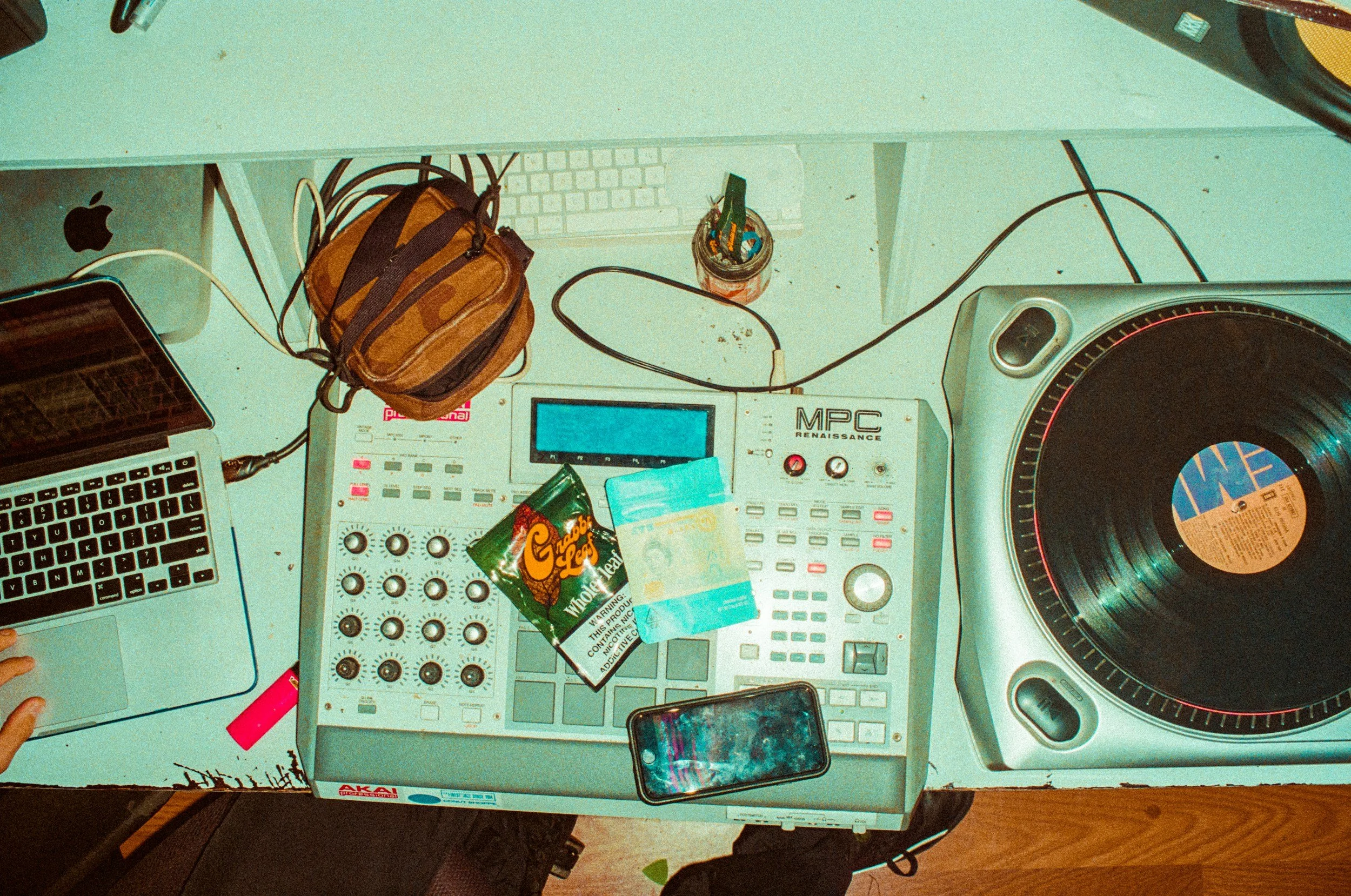 A cluttered DJ workspace with a laptop, mixing controller, turntable, small bag, snacks, and various cords on a white table.