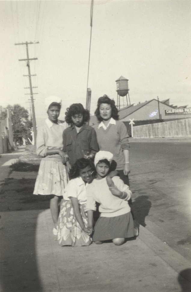 Five women standing and kneeling on a sidewalk in front of a building with a water tower in the background. The women are dressed in vintage clothing.