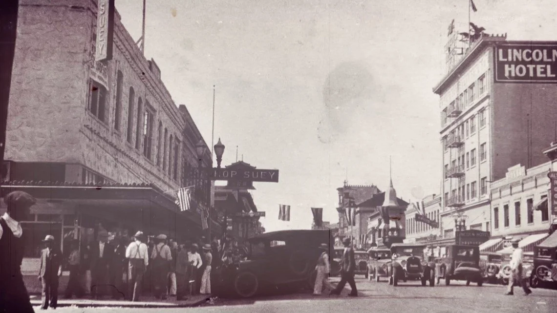 Historical black-and-white photo of a busy downtown street with pedestrians, vintage cars, and storefronts, including the Lincoln Hotel and Top Suey restaurant.