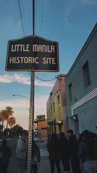 Street scene with a sign that reads 'Little Manila Historic Site,' people walking along the sidewalk, and colorful buildings under a blue sky.