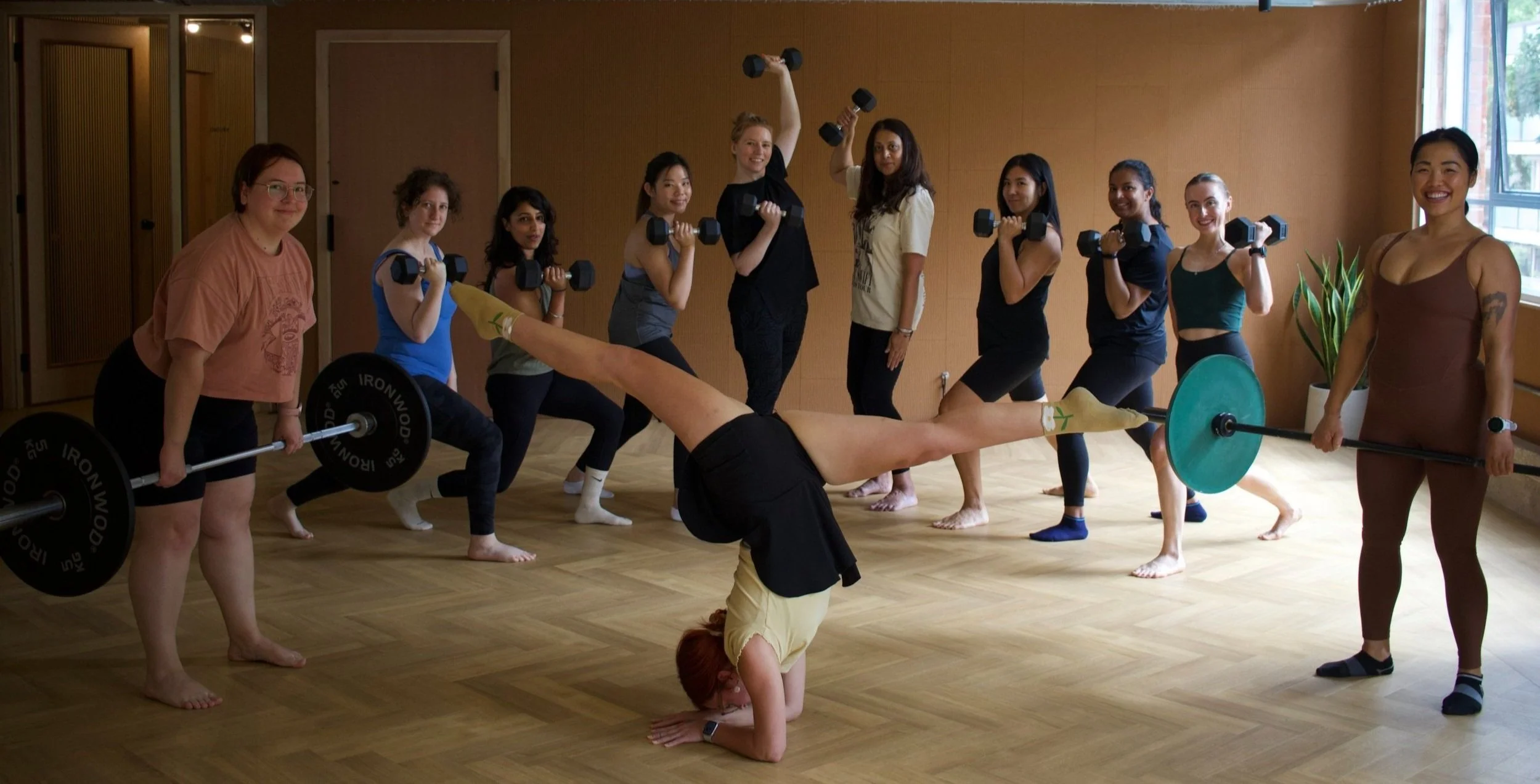 A group fitness class with women lifting dumbbells, doing squats, and a woman performing a headstand while others smile and participate inside a brightly lit room.