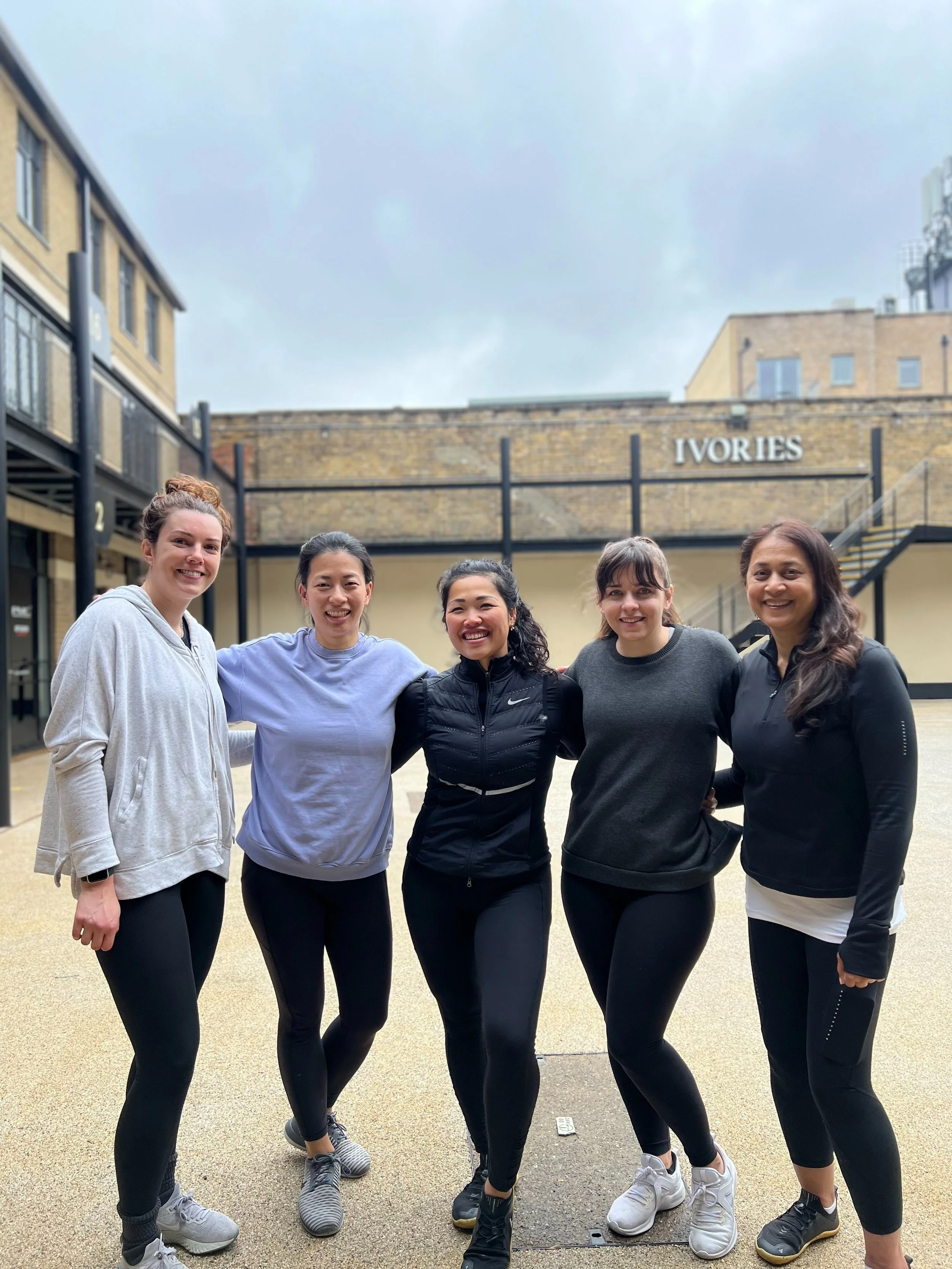 Group of five women dressed in athletic clothing standing together outdoors in an open courtyard, smiling at the camera, with a building labeled "IVORIES" in the background.