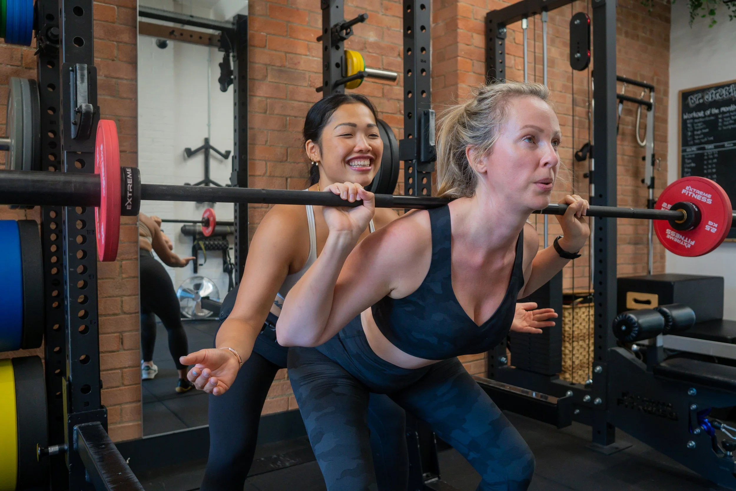 Two women exercising in a gym, one is doing a barbell squat while the other assists.