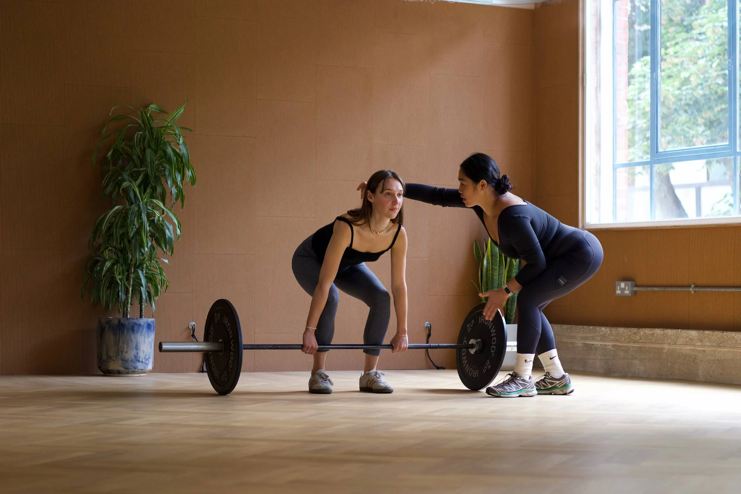 A woman in workout clothes is lifting a barbell with weights on both ends, while a trainer assists her by giving instructions and support during her exercise in a gym with wooden floors, a window, and plants.