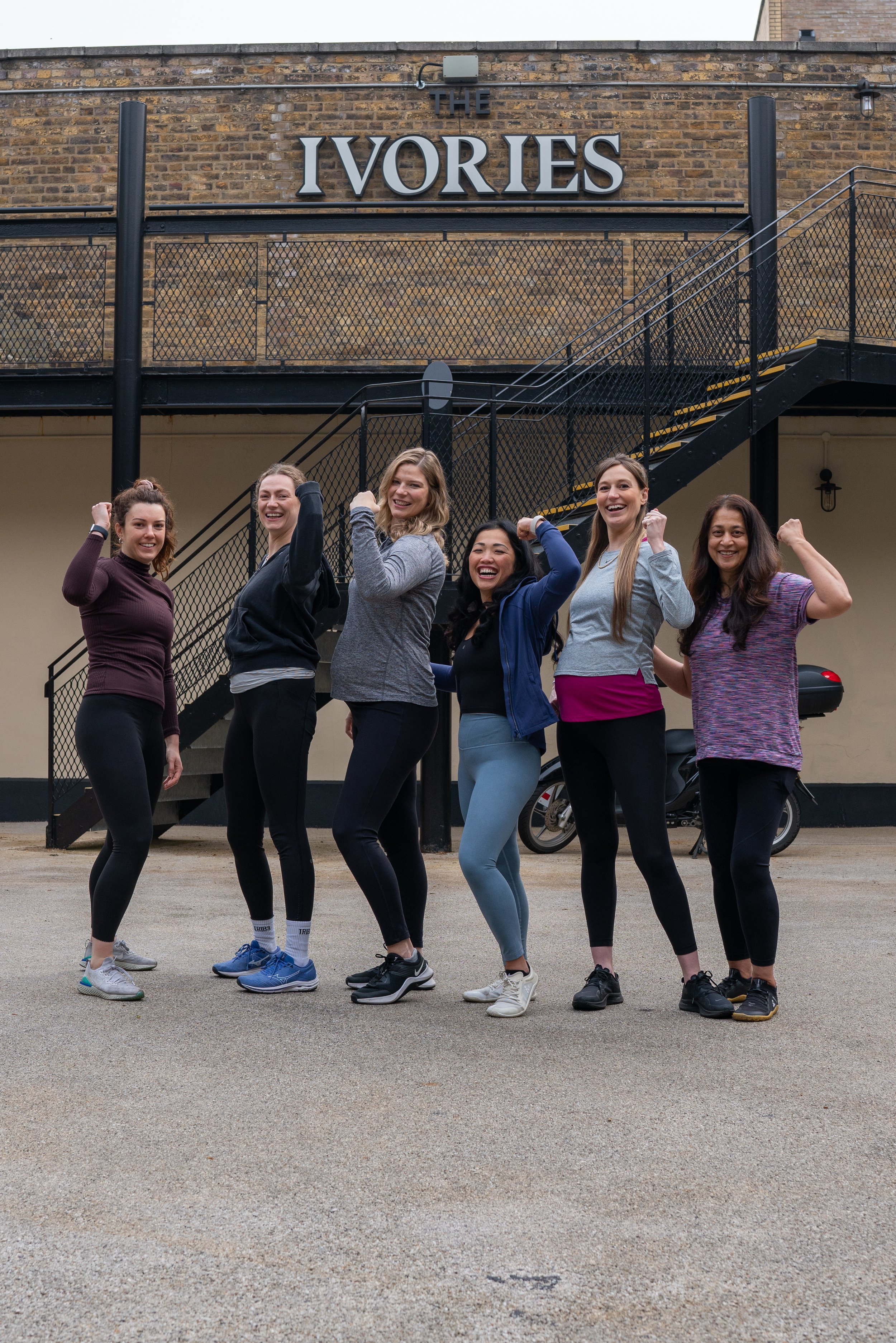 Six women in workout clothes smiling and flexing their arms in front of a brick building with a sign that reads "The Ivories".