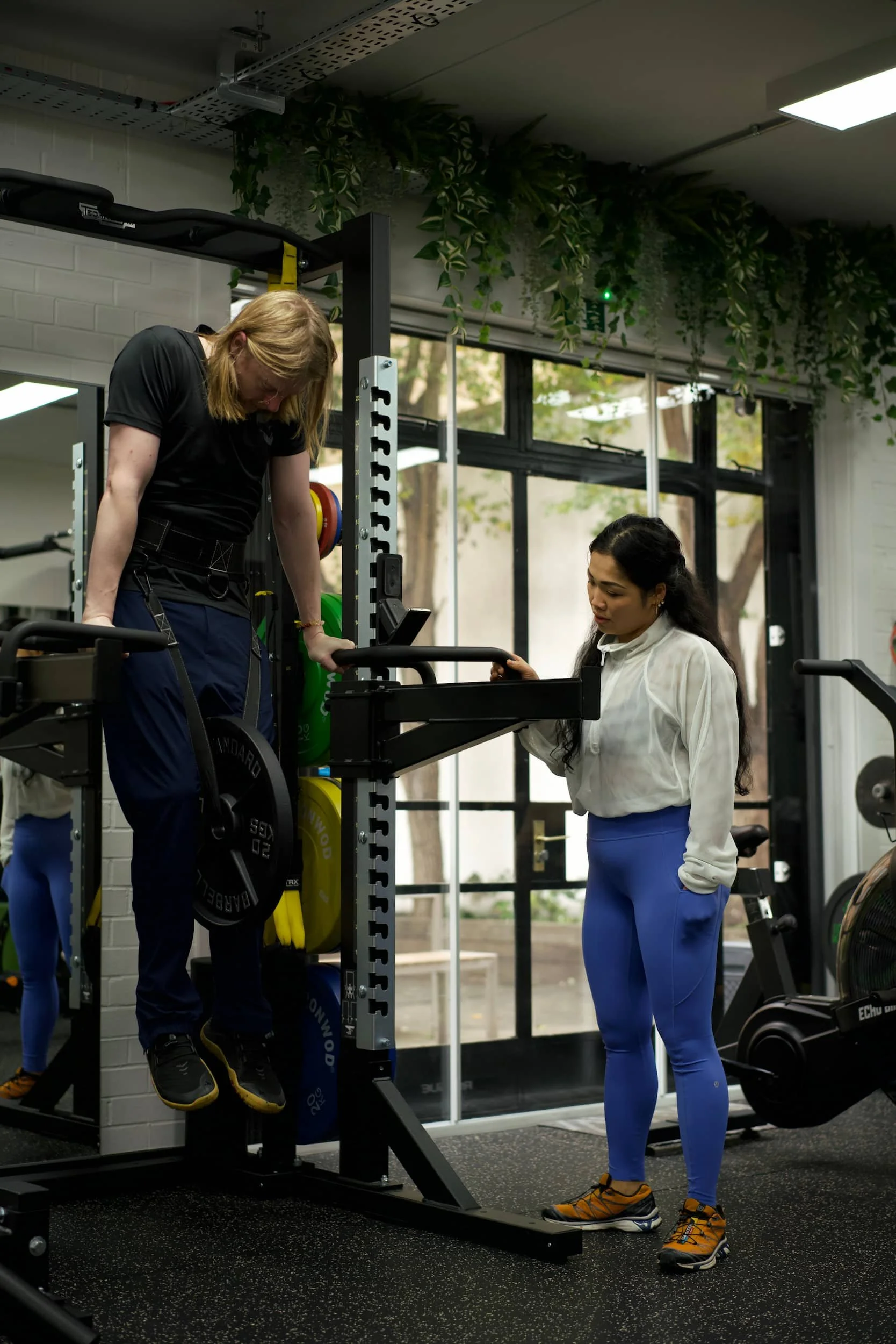 A woman receiving guidance from a trainer during a workout in a gym, with weights and exercise equipment visible.