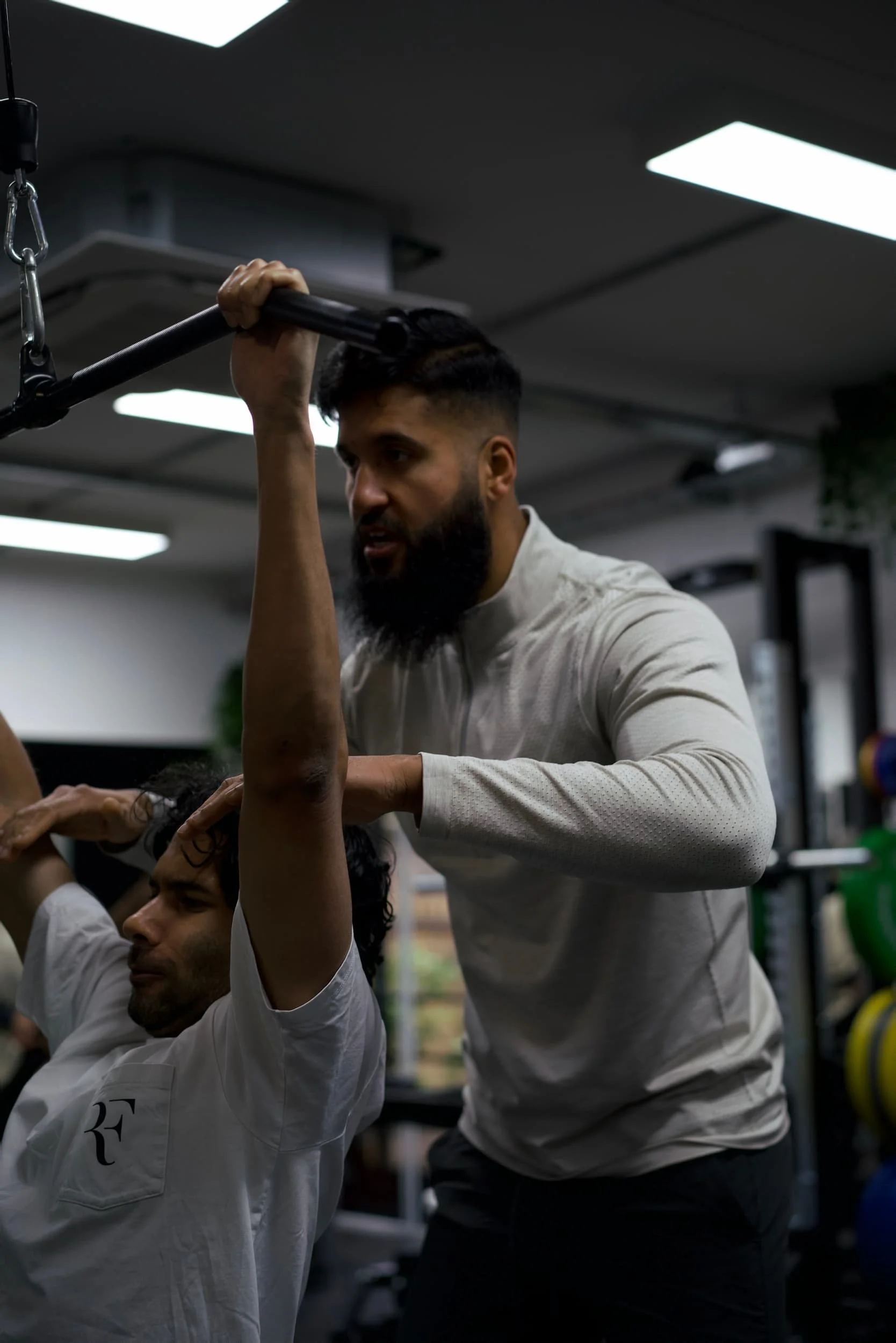 Personal trainer assisting a man during a workout at the gym, with weightlifting equipment in the background.