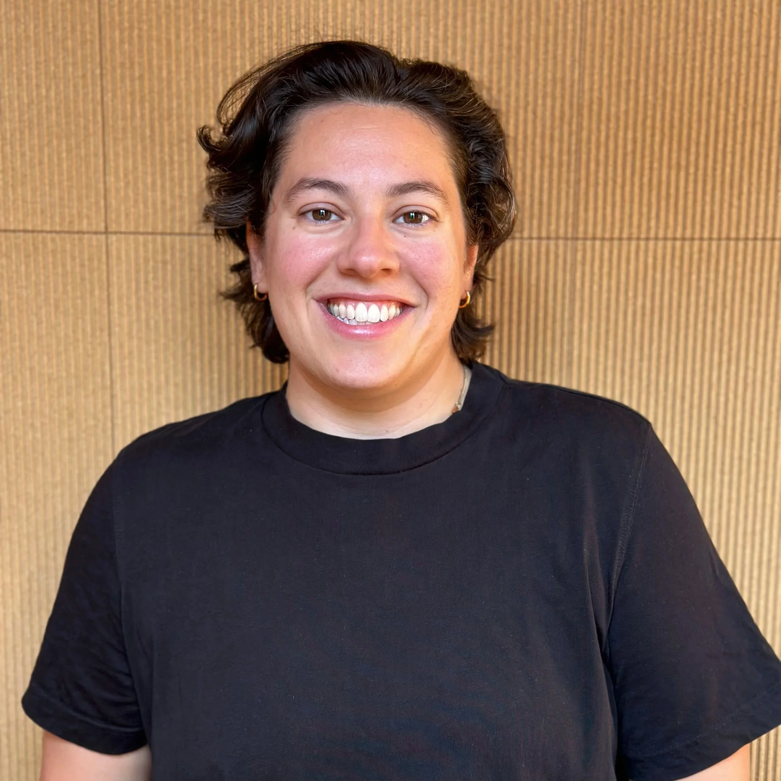 A woman with short dark hair, wearing a black t-shirt and small gold hoop earrings, smiling in front of a wooden wall.