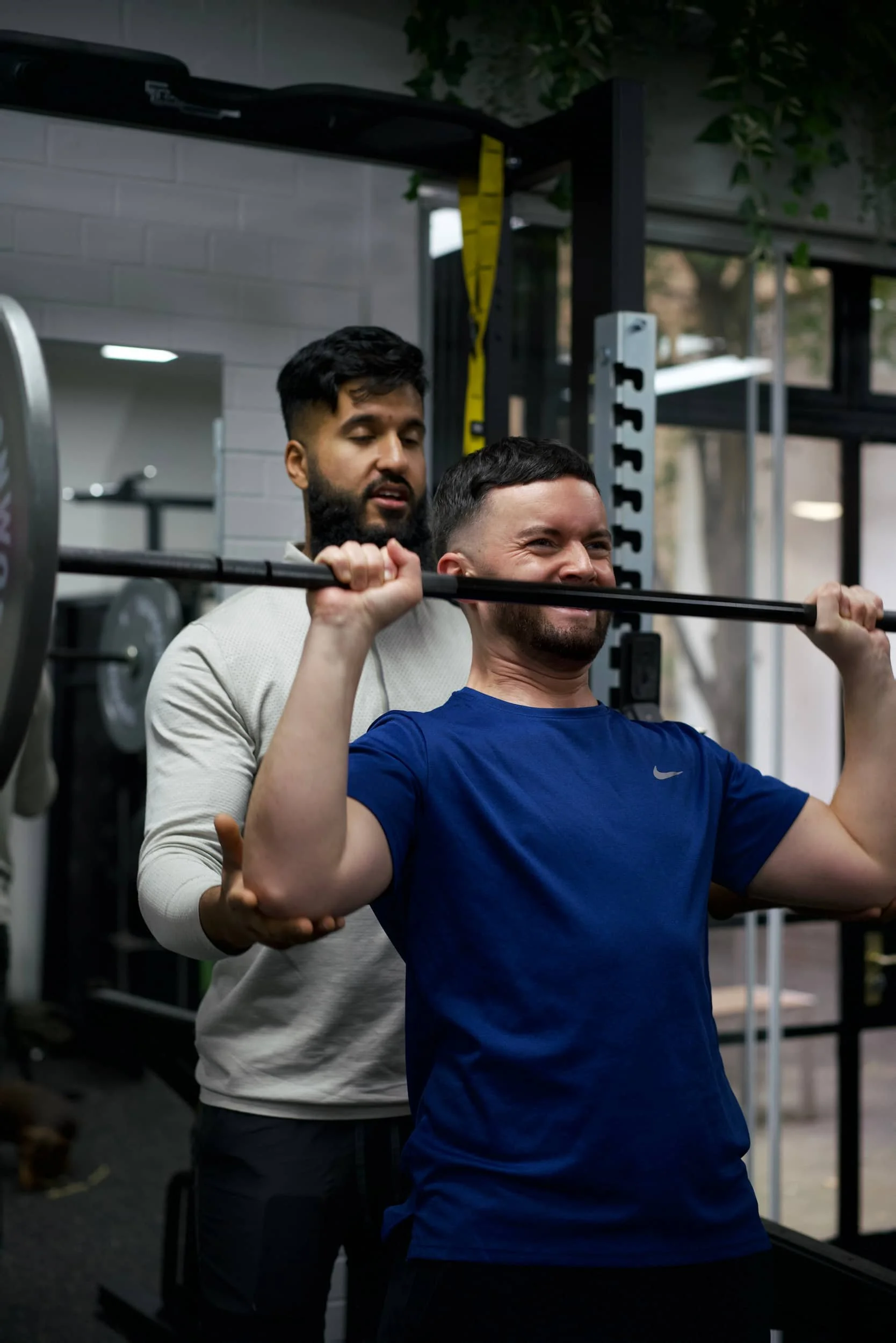 A man in a blue shirt is lifting a barbell with weights, assisted by a trainer in a gym.
