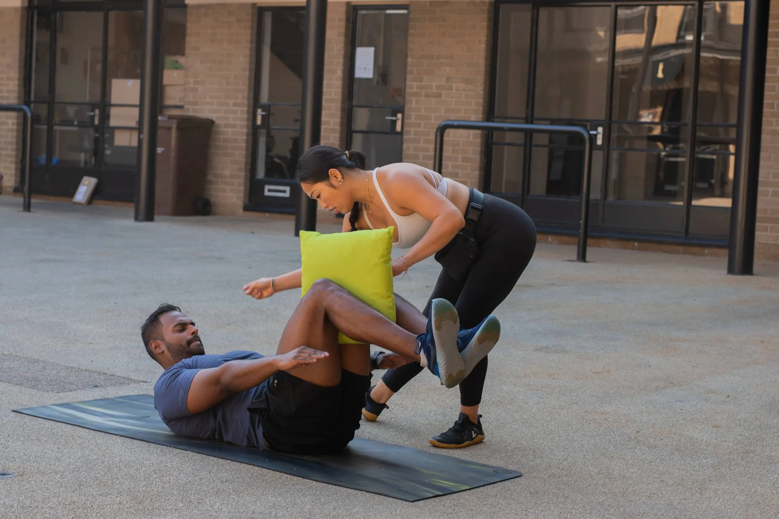 A woman helping a man during outdoor exercise, with the man lying on a yoga mat and holding a green cushion while the woman is leaning over him.