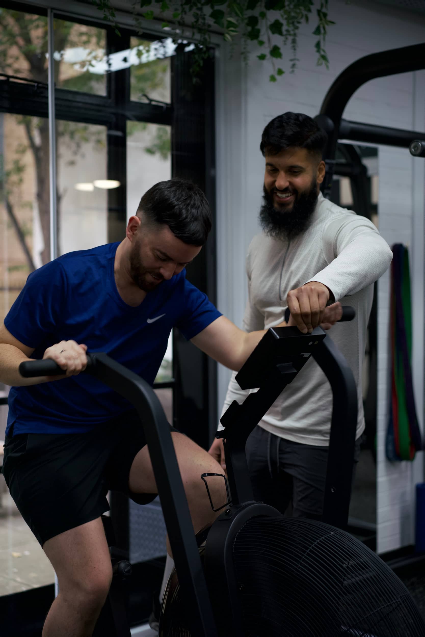 Two men are working out with a rowing machine in a gym. One man is using the machine while the other is coaching or assisting him, both smiling.