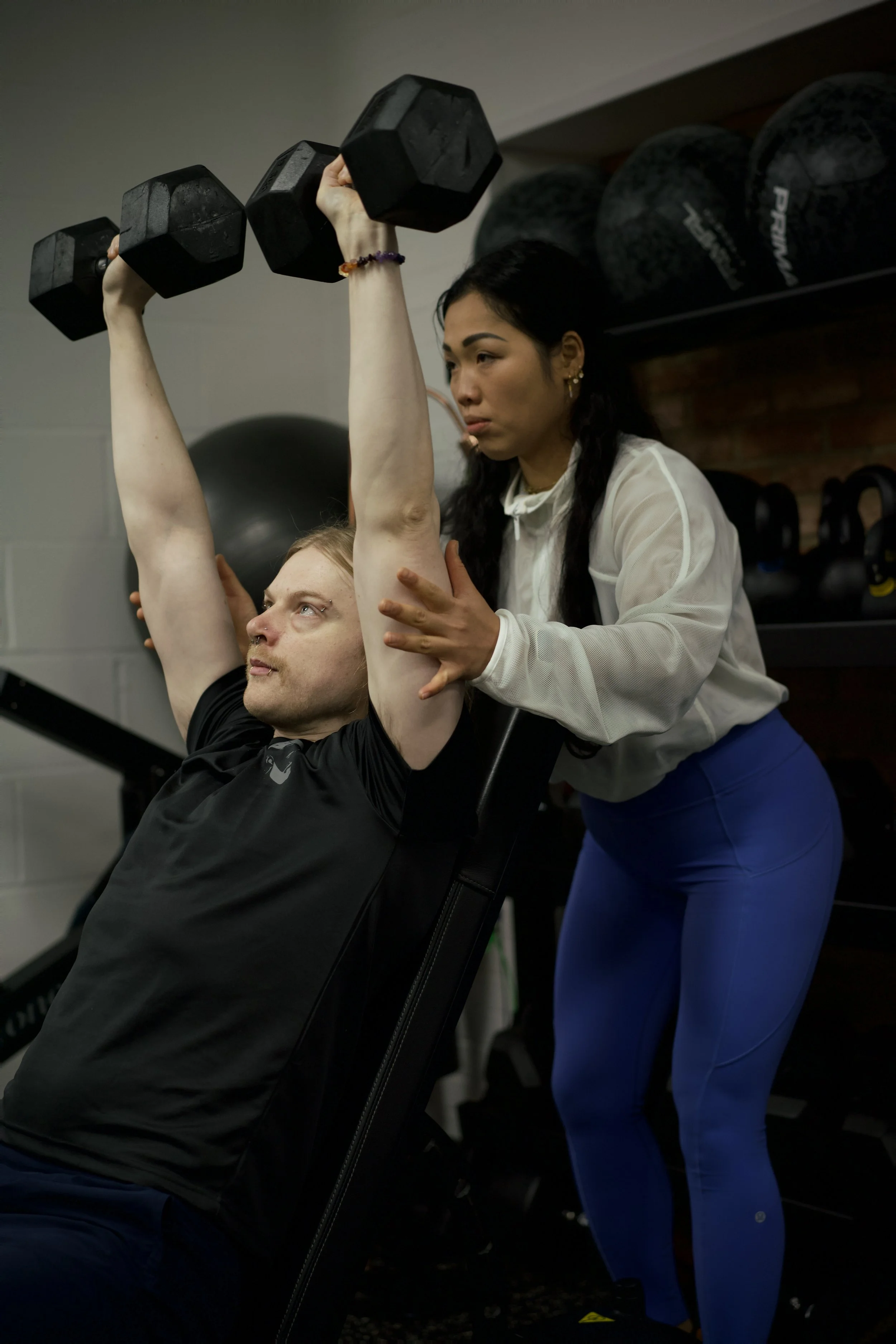 A woman in blue leggings and a loose white top spotters a man in a black t-shirt doing a shoulder press with black dumbbells in a gym, with her hand supporting his back.
