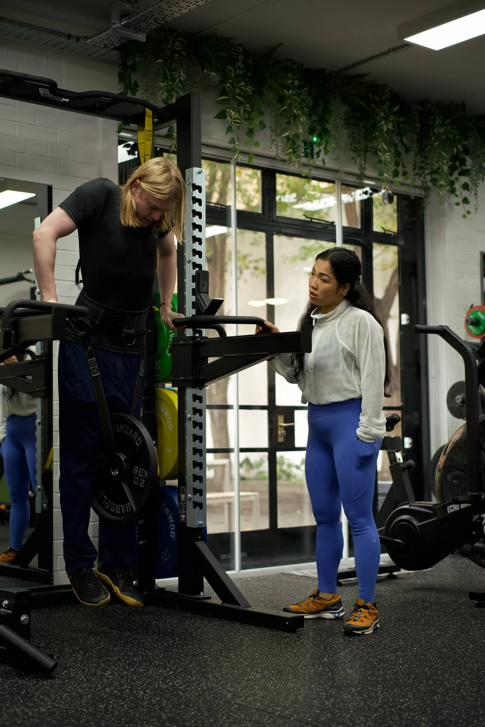 A fitness coach assisting a woman with a workout on a stair stepper machine in a gym.