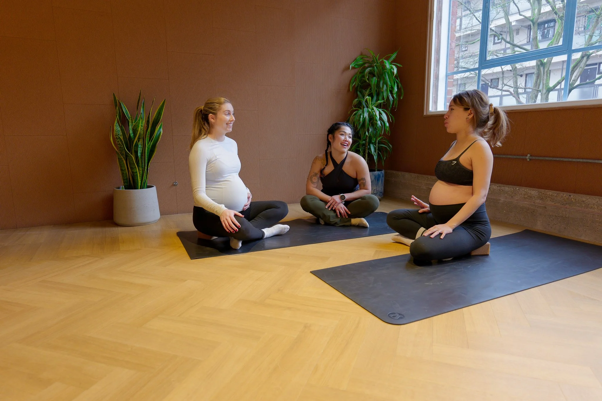 Three women in yoga clothes sitting on yoga mats in a room with wooden flooring and brown walls, engaging in conversation during a prenatal yoga class.