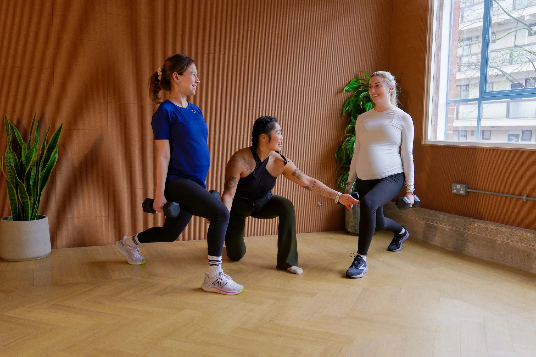 Three women exercising indoors with dumbbells, one of them is holding a dumbbell in each hand while the other two are holding one dumbbell each, smiling and engaging with each other, in a room with a wooden wall, plants, and a large window.