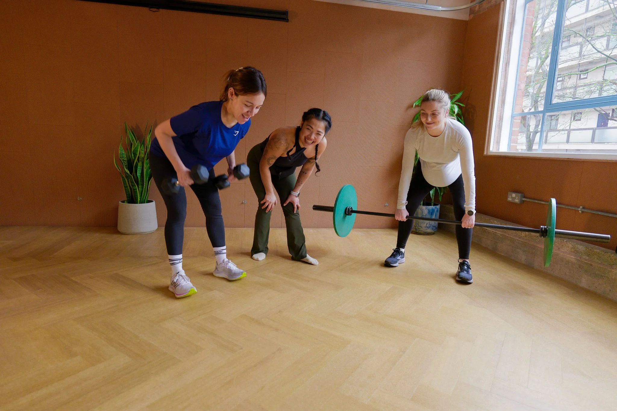 Three women in workout attire exercising in a studio, with one lifting dumbbells, another holding a barbell, and the third, who is built into the session, smiling and leaning forward. The room has wooden floors, potted plants, and a large window.
