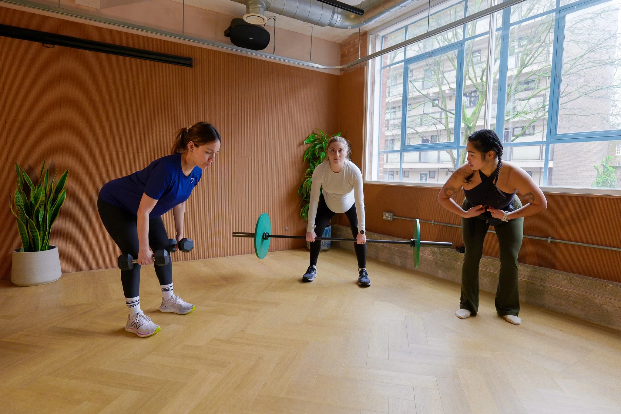 Three women in workout clothes lifting weights and preparing for exercise in a bright gym room with large windows, hardwood floors, and potted plants.