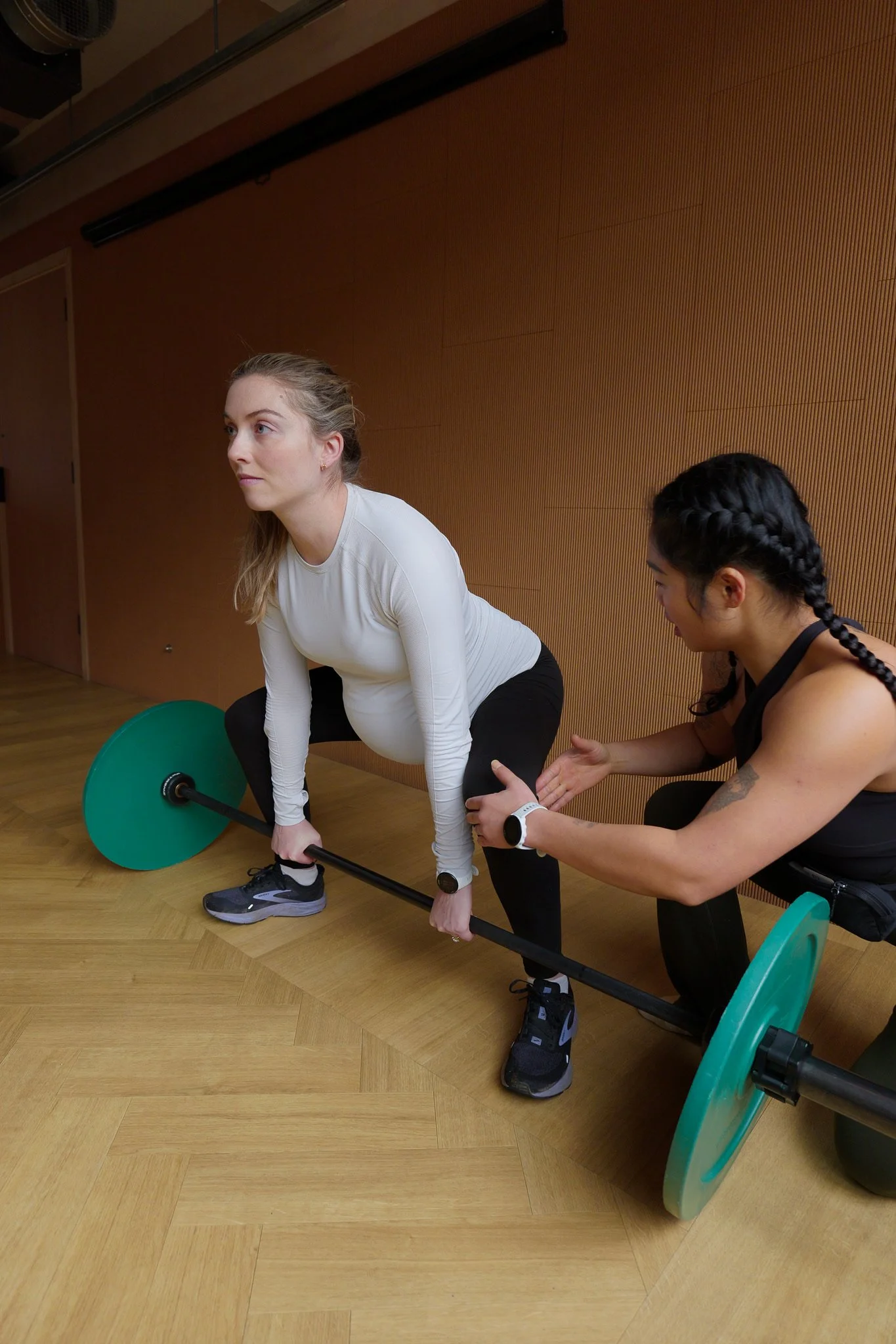 A pregnant woman lifting a barbell with guidance from her trainer during a workout session in a gym.