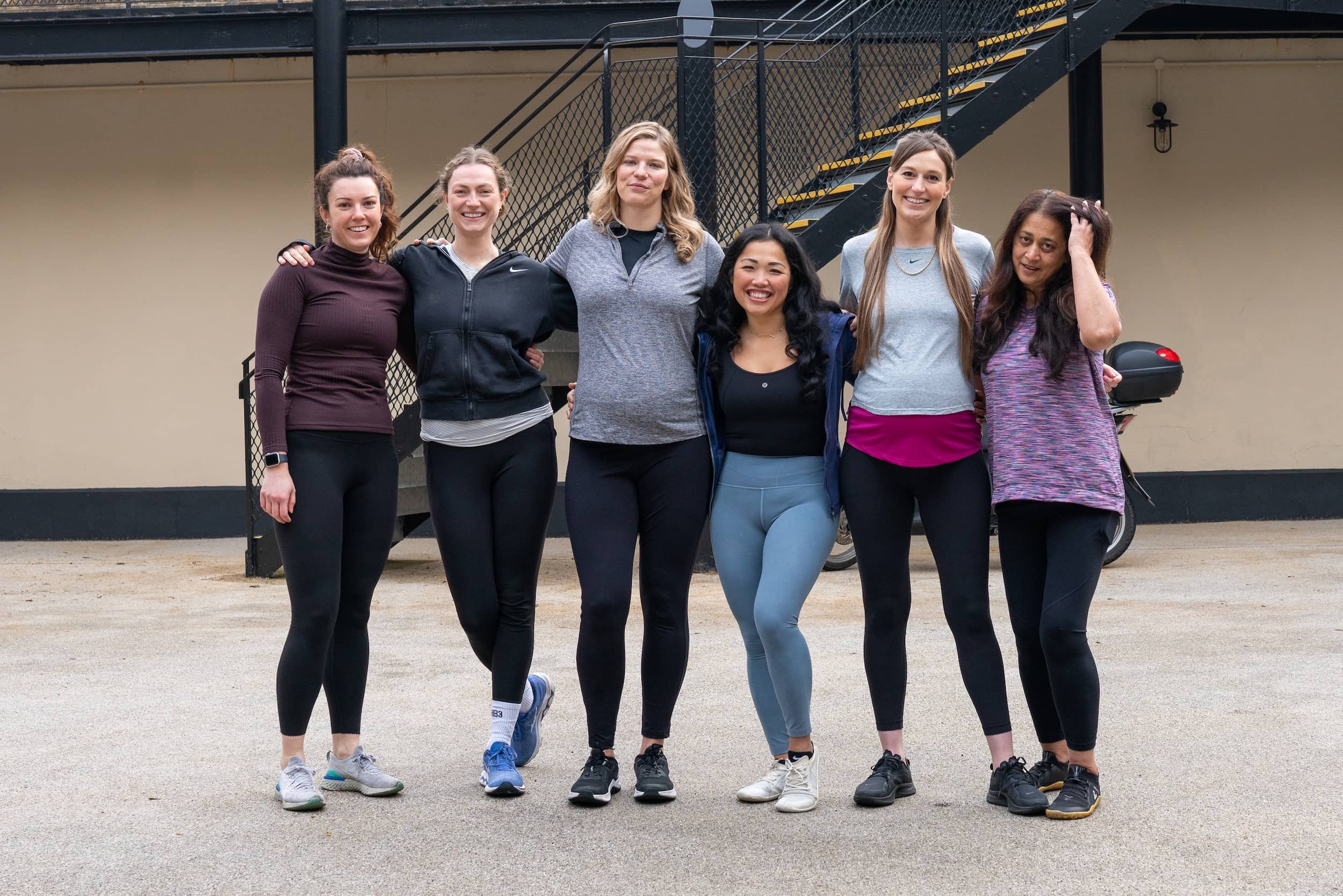 Six women standing outdoors, arms around each other, smiling, dressed in athletic wear.