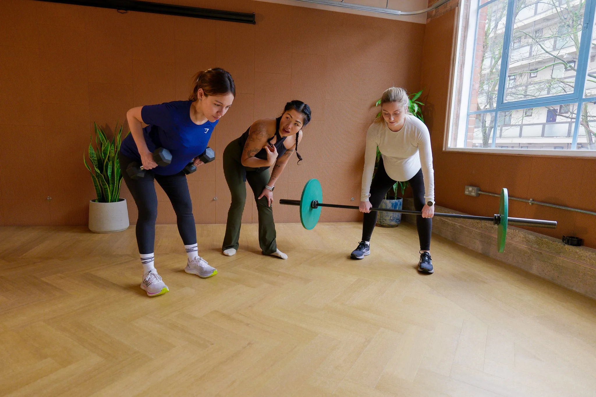 Three women in workout clothes practicing weights and strength training indoors, with one woman coaching or instructing, in a room with wooden floors and walls, and large windows.