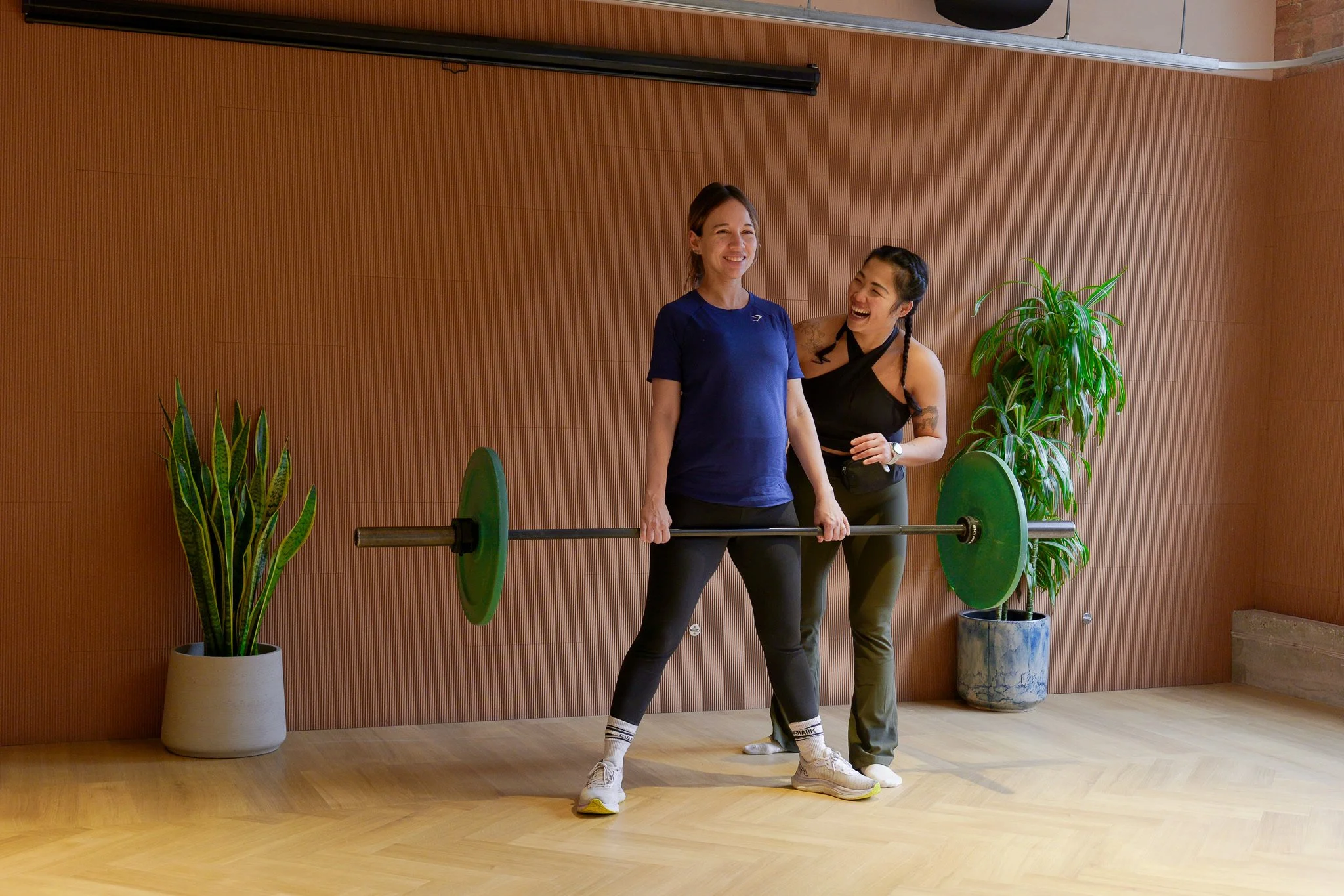 Two women laughing, one lifting a barbell in a fitness studio with potted plants and a wooden floor.