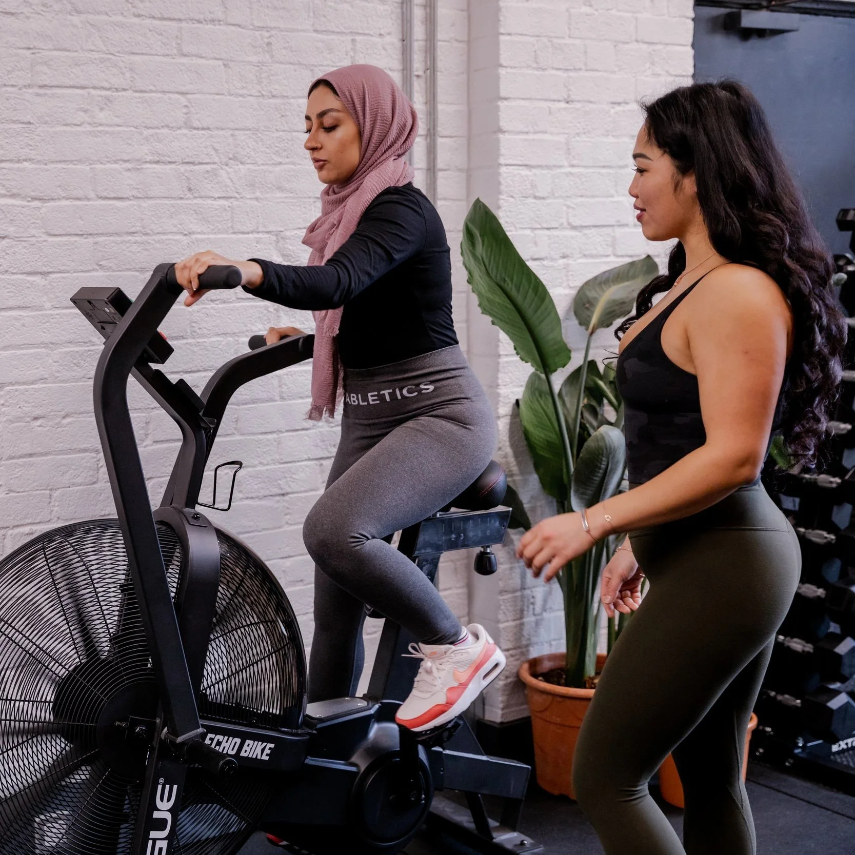 A woman in a hijab is using a stationary exercise bike while another woman, standing beside her, observes or supervises. The scene is inside a gym with a white brick wall and some large green plants in the background.