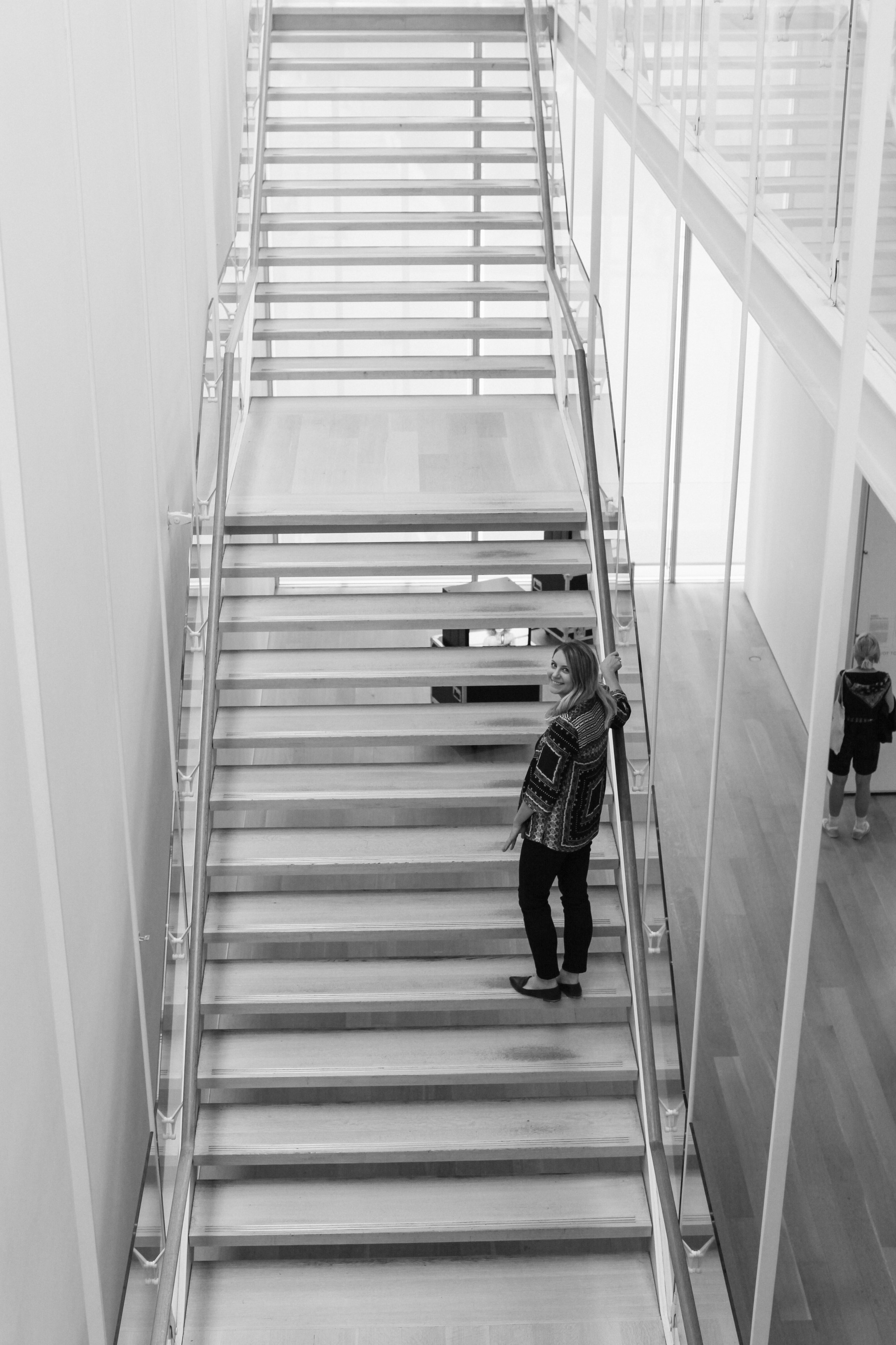 A woman standing on a wooden staircase in a modern building, smiling at the camera. Two children are at the bottom of the stairs, one holding onto the railing.