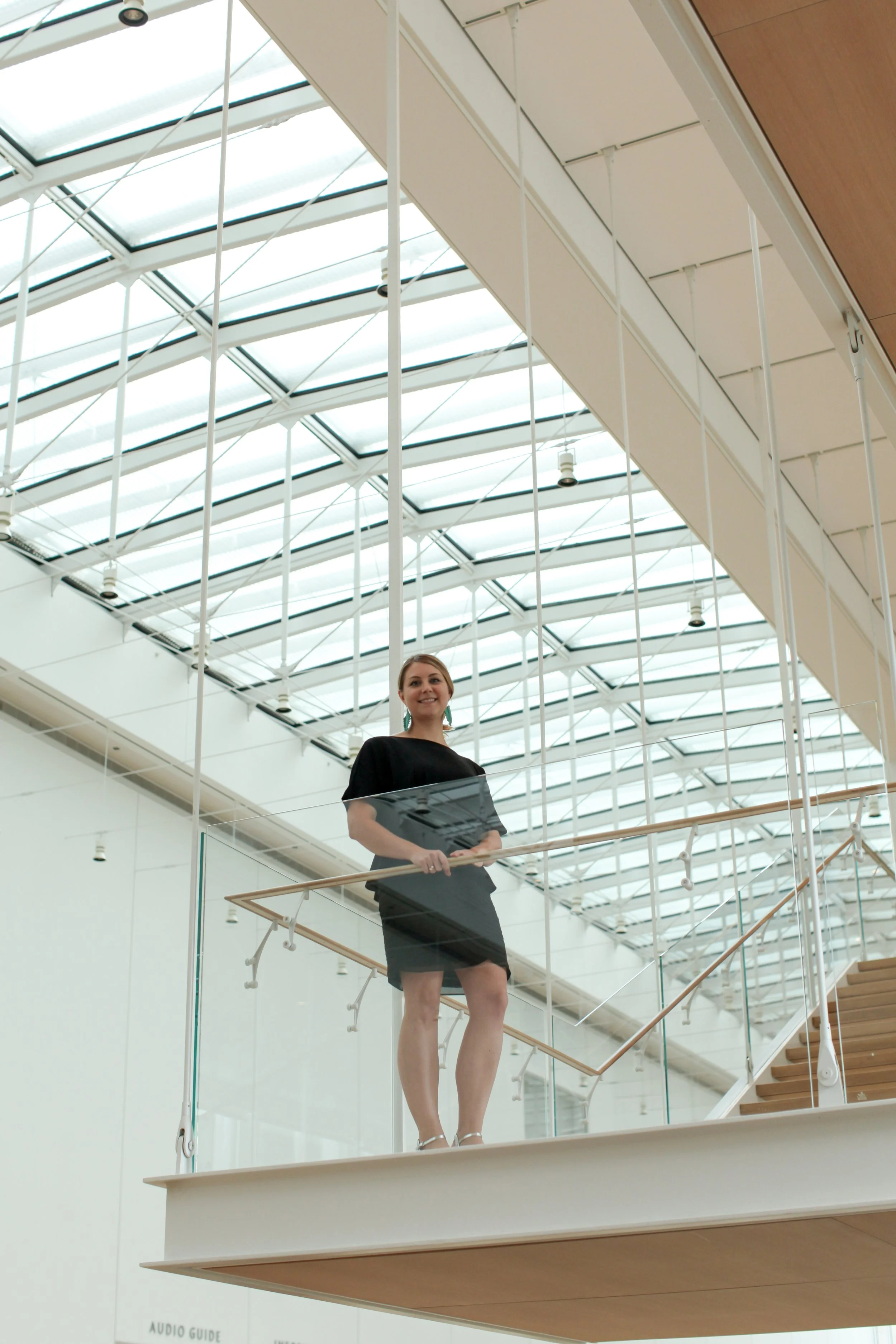 A woman in a black dress holding a black folder or tablet, standing on a glass balcony inside a modern building with a glass ceiling.