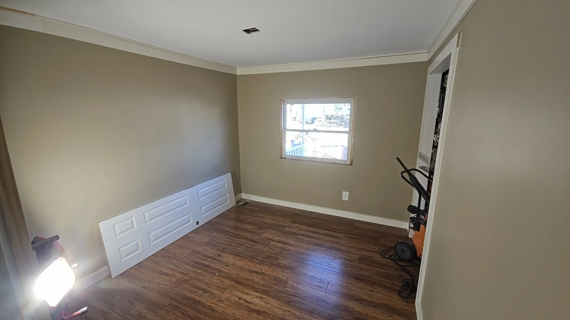 Empty room with hardwood flooring, beige walls, an unfinished window, recessed lighting, a white door panel on the wall, and a vacuum cleaner in the corner.