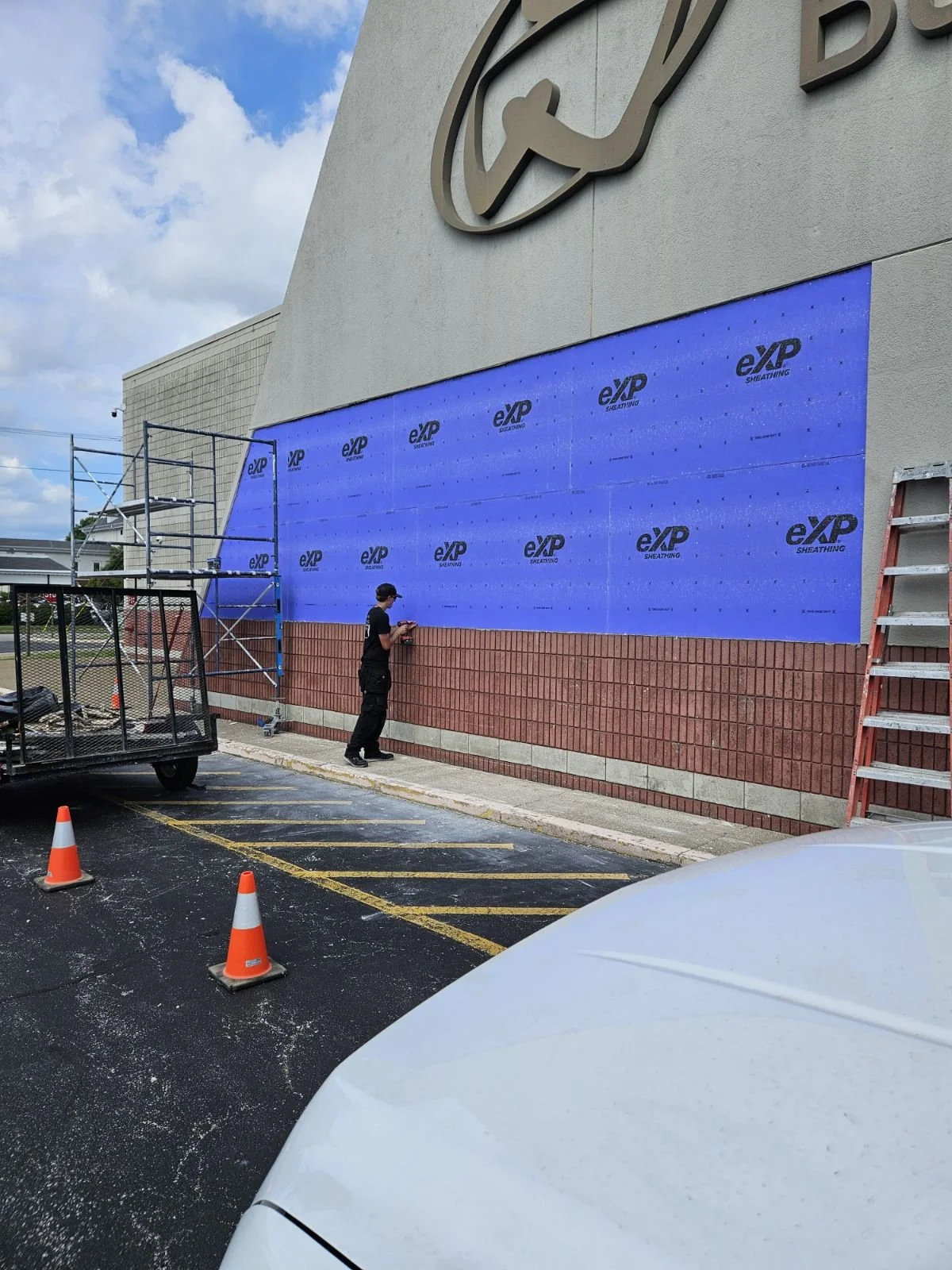 A worker is installing blue sheathing material on the lower part of a building wall, with scaffolding and tools nearby, in a parking lot with pending construction.
