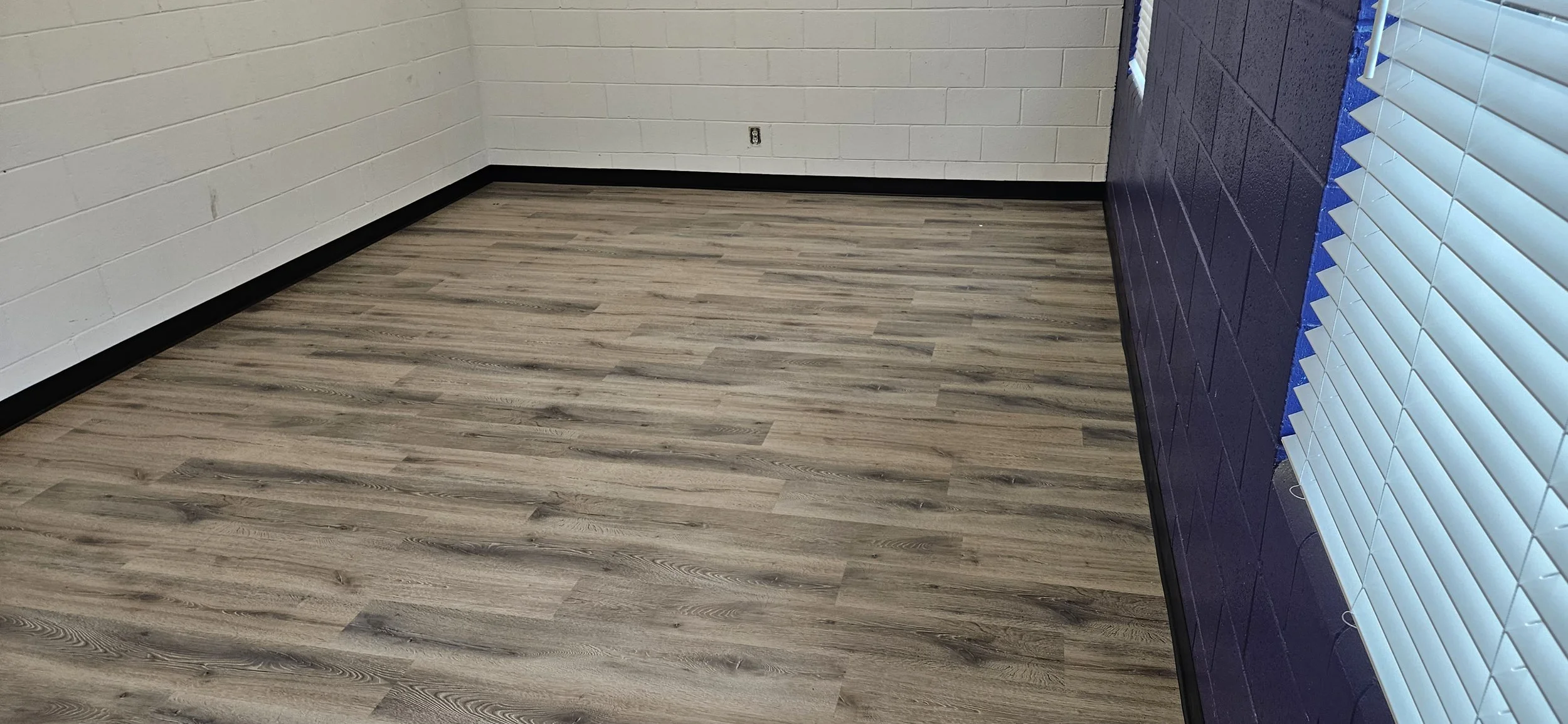 Empty room with white brick walls, grayish wood flooring, a purple wall, and windows with white blinds.