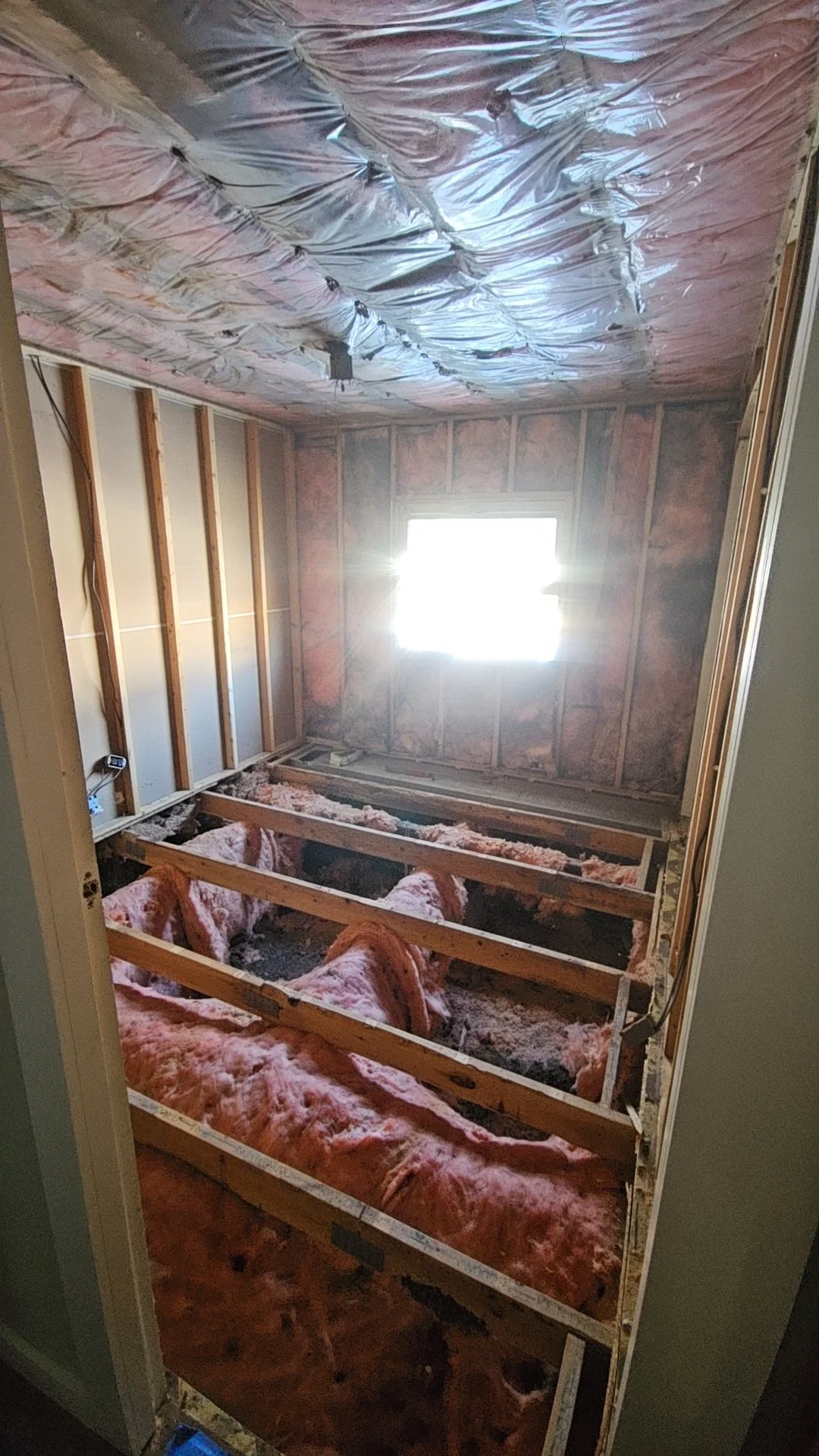 Room under construction with exposed wooden framing, pink insulation, and a window letting in sunlight.